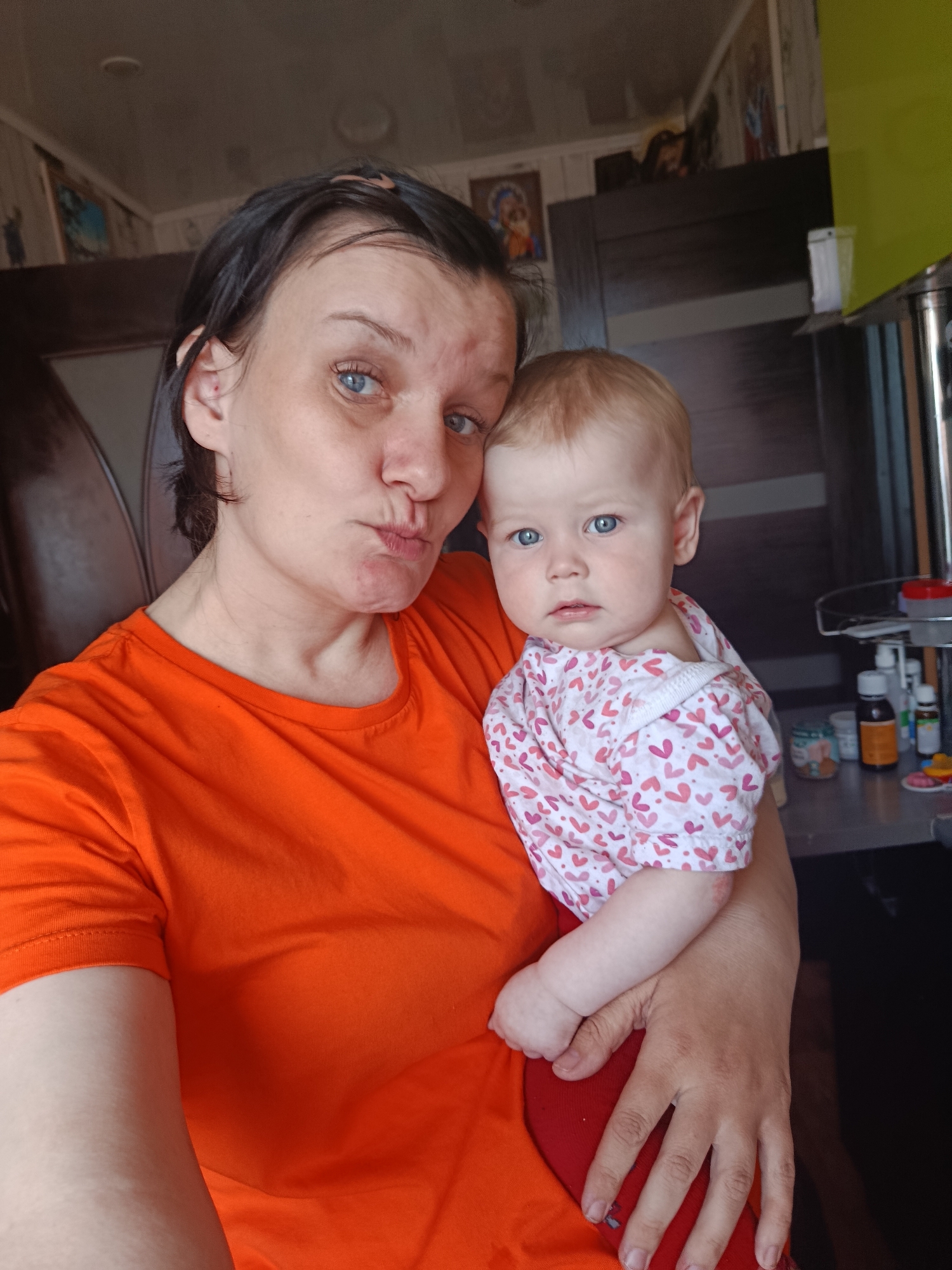 A woman with short dark hair, wearing an orange t-shirt, is holding a baby with blonde hair and blue eyes, wearing a pink and white heart-patterned shirt and red pants. The woman has her lips pursed in a kissing gesture. The background includes a dark brown wooden cabinet and a countertop with various items on it, including bottles and containers. The wallpaper is patterned with vertical stripes and small images, which are not distinctly visible. The photograph appears to be taken indoors. There is no specific evidence that the image was taken in Фоки, Russia.