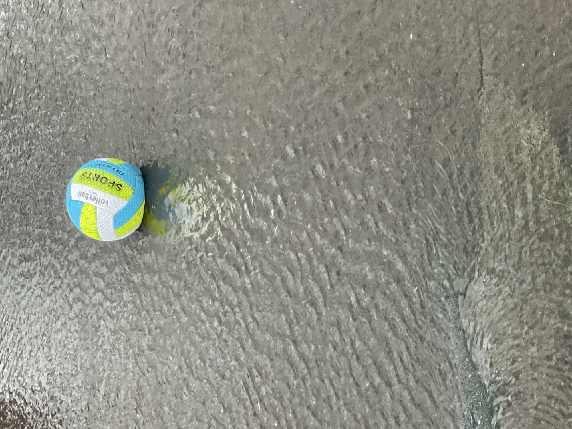 A volleyball with blue, white, and yellow panels sits on a wet, dark gray surface with rippling textures. Text on the ball reads "Volleyball," "SPORTS," and "OFFICIAL." The surface appears to be asphalt or a similar material covered in a thin layer of water, possibly from rain or a puddle, reflecting the ball and the sky. The overall scene is outdoors. The location is Khimki, Russia.