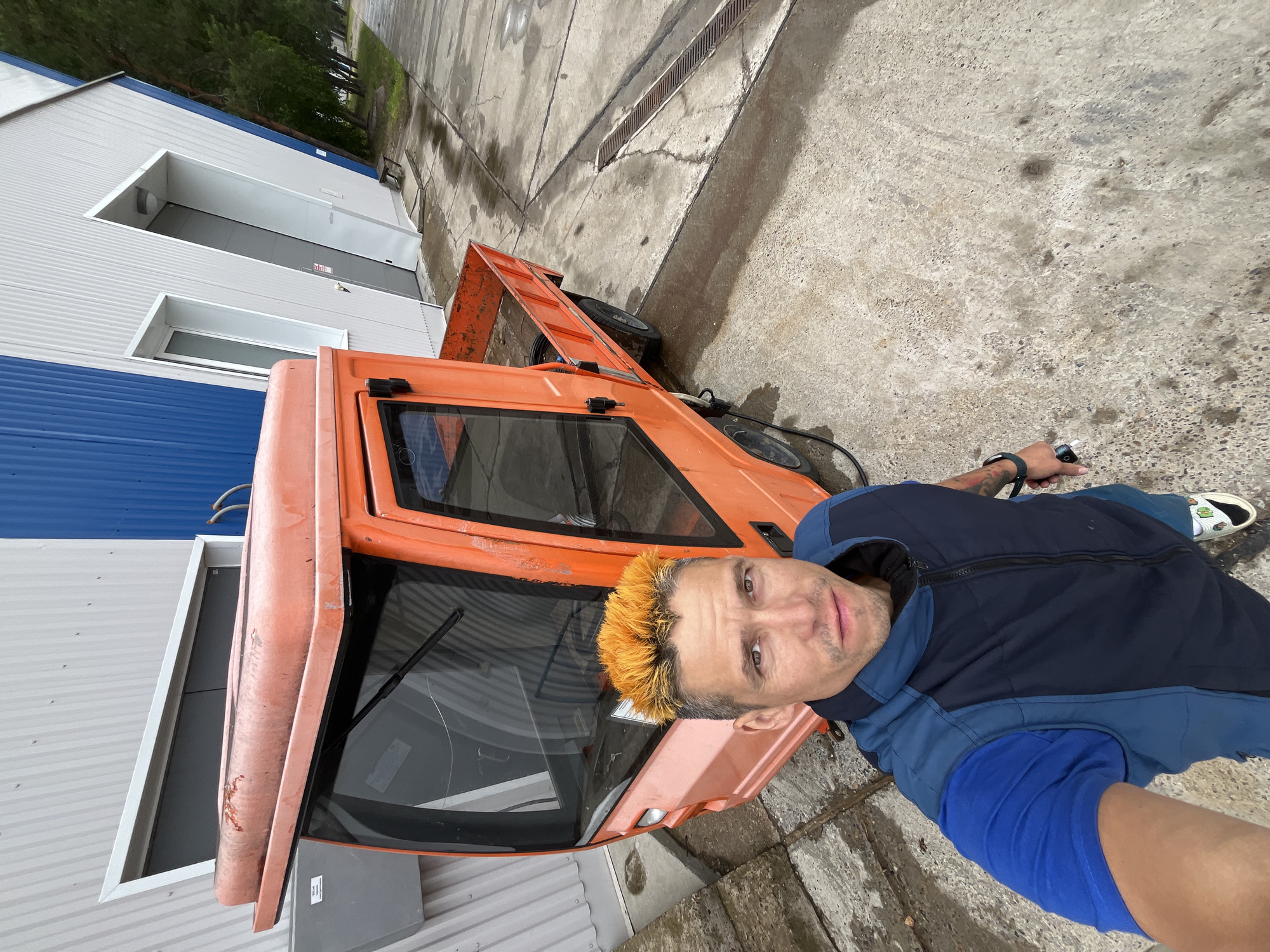 An over-the-shoulder selfie is captured by a man with bright orange hair, standing next to an orange utility vehicle. The man is wearing a blue t-shirt and a dark blue vest. The utility vehicle has a closed cabin and an open flatbed in the back. It is parked on a wet concrete surface. Behind the vehicle is a modern building with grey and blue corrugated metal siding and large garage doors. The scene appears to be an industrial or service area. The background shows trees and a sidewalk. The image is taken from a high angle.