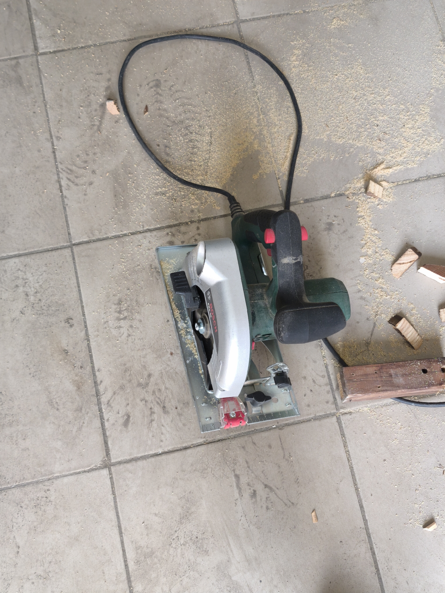The image shows a circular saw on a tiled floor, surrounded by sawdust and small pieces of wood. The saw is primarily silver and green, with a black handle and a red safety switch. A black power cord extends from the saw. The floor is composed of square tiles, and the scene appears to be indoors. The presence of the saw, wood, and sawdust suggests woodworking activity. The location cannot be determined to be in Moscow, Russia from this image.