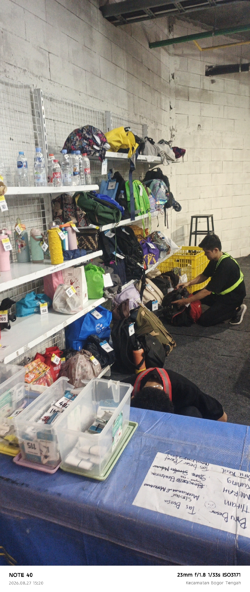 The image depicts an indoor storage area, possibly a warehouse or event venue, filled with numerous items. On white shelves, various objects are displayed, including water bottles, patterned bags, and brightly colored water bottles. The shelves are constructed of metal wire grids. On the floor, the scene is cluttered with bags of different sizes and colors, backpacks, and a yellow plastic crate. A man in a black shirt with a reflective yellow vest is crouching on the floor, interacting with a black and red backpack. In the foreground, two clear plastic containers are placed on a blue tarp-covered surface. One of these containers holds smaller items. Another person, wearing a black shirt with a reflective red vest, is lying face down on the blue tarp, their head resting on their arms. Text on a white paper attached to the blue tarp is partially visible, appearing to be a list of items with descriptions in Indonesian, suggesting an inventory or labeling system. The wall in the background is made of light-colored concrete blocks. Above the shelves, metal conduits and pipes are visible. The lighting suggests an artificial indoor environment. The timestamp at the bottom left indicates the photo was taken on August 27, 2026, at 15:20. The text "Kecamatan Bogor Tengah" suggests the location is within the Central Bogor District, Bogor City, Indonesia.