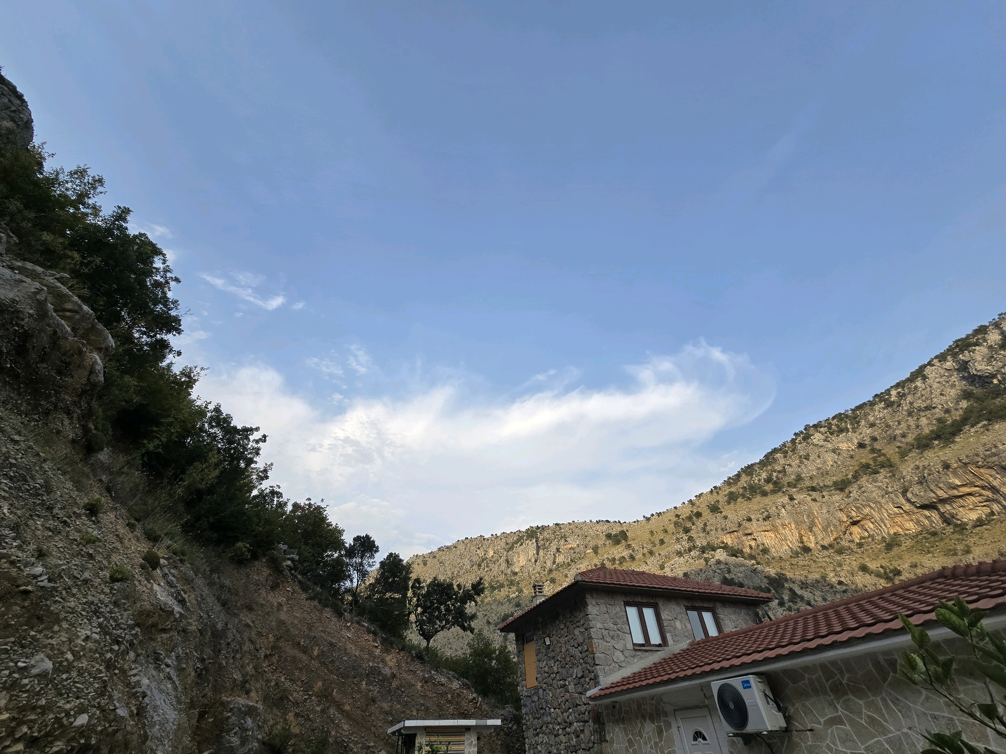 The image depicts a mountainous landscape with a partially constructed stone building in the foreground, located in the city of Podgorica, Montenegro. The sky is a clear blue with scattered white clouds. To the left, a steep, rocky slope is covered with sparse vegetation and trees. To the right, a substantial mountain range rises, characterized by rugged rock formations and scattered greenery. The building itself is constructed from rough-hewn stones and features a red-tiled roof. One section of the building has visible windows with brown frames. An outdoor air conditioning unit is mounted on the exterior wall of another part of the building, which appears to be under construction or renovation. The overall scene suggests a remote, rural setting within or near Podgorica.