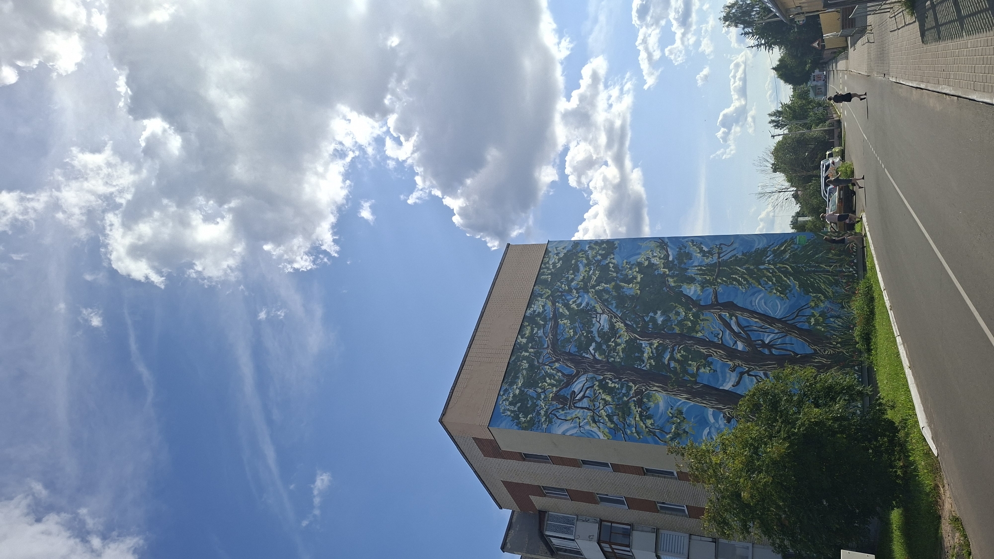 A vertical, eye-level, long shot captures a mural of trees on the side of a multi-story apartment building, located on a street in the city of Myadzyel, Belarus. The building, predominantly light brown brick with a beige trim, features a balcony on its left side. The mural, which covers a significant portion of the building's facade, depicts large, gnarled trees with green foliage against a bright blue sky. To the right of the building, a paved road runs parallel to a sidewalk lined with shops and houses. Several people are visible on the street. Two individuals are walking down the road, while another is standing near a car. Further down the street, a person in a black dress is walking towards the camera. The sky is a vibrant blue with scattered white clouds, and sunlight casts shadows on the scene.