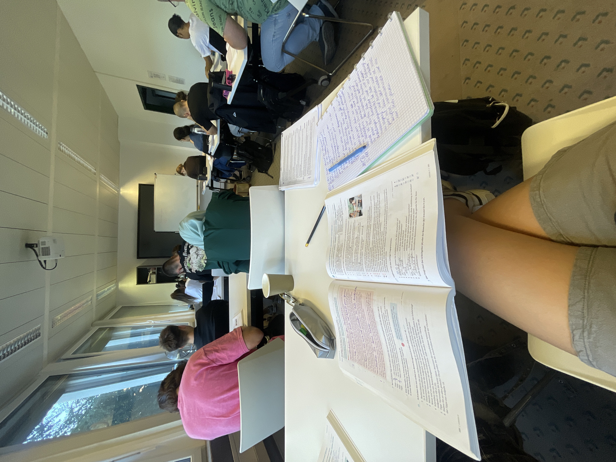The image shows a classroom in Solingen, Germany, with students sitting at desks. In the foreground, a person's legs and lap are visible, with an open textbook and notebook on the desk in front of them. The textbook is open to a page with German text and exercises. A blue pen rests on the notebook, which is filled with handwritten notes. The classroom is brightly lit, with large windows on the left side offering a view of trees and a cloudy sky. Other students are visible in the background, facing forward towards a whiteboard or screen that is not fully in view. The environment appears to be a typical educational setting.