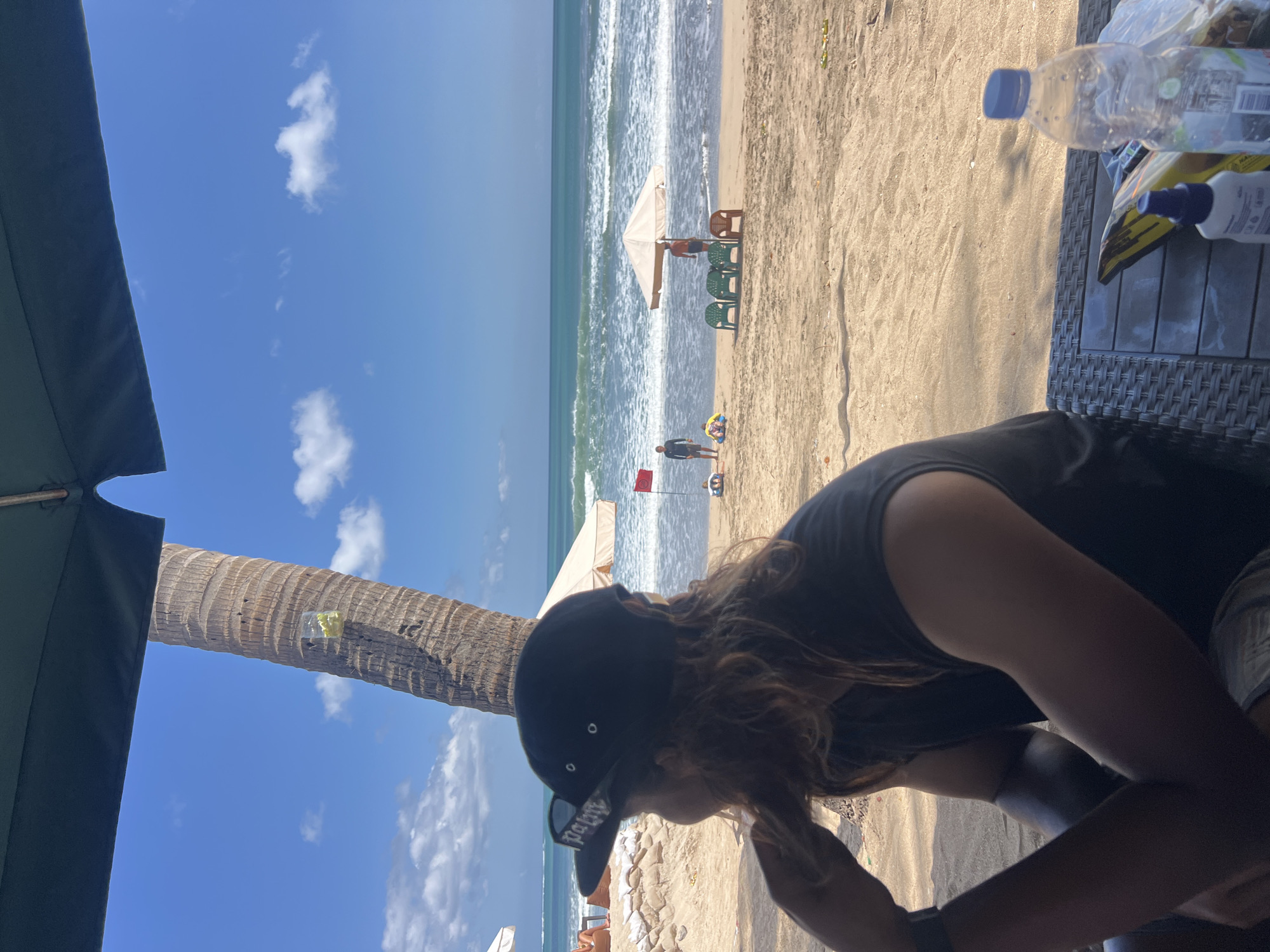In Kuta, Indonesia, a person wearing a black t-shirt and a black baseball cap with white lettering sits at a table on a sandy beach, facing away from the camera and looking out at the ocean. The person has long, wavy brown hair and a visible tattoo on their left arm. In the foreground, a plastic bottle of water and a white bottle with a blue cap are on the table. Beyond the person, the beach is dotted with beach umbrellas, lounge chairs, and a few people. One person stands near a red flag, while others are seated or playing on the sand. The ocean has moderate waves, and the sky is a clear blue with a few scattered clouds. A palm tree is visible to the left of the frame, partially obscured by a dark green umbrella.
