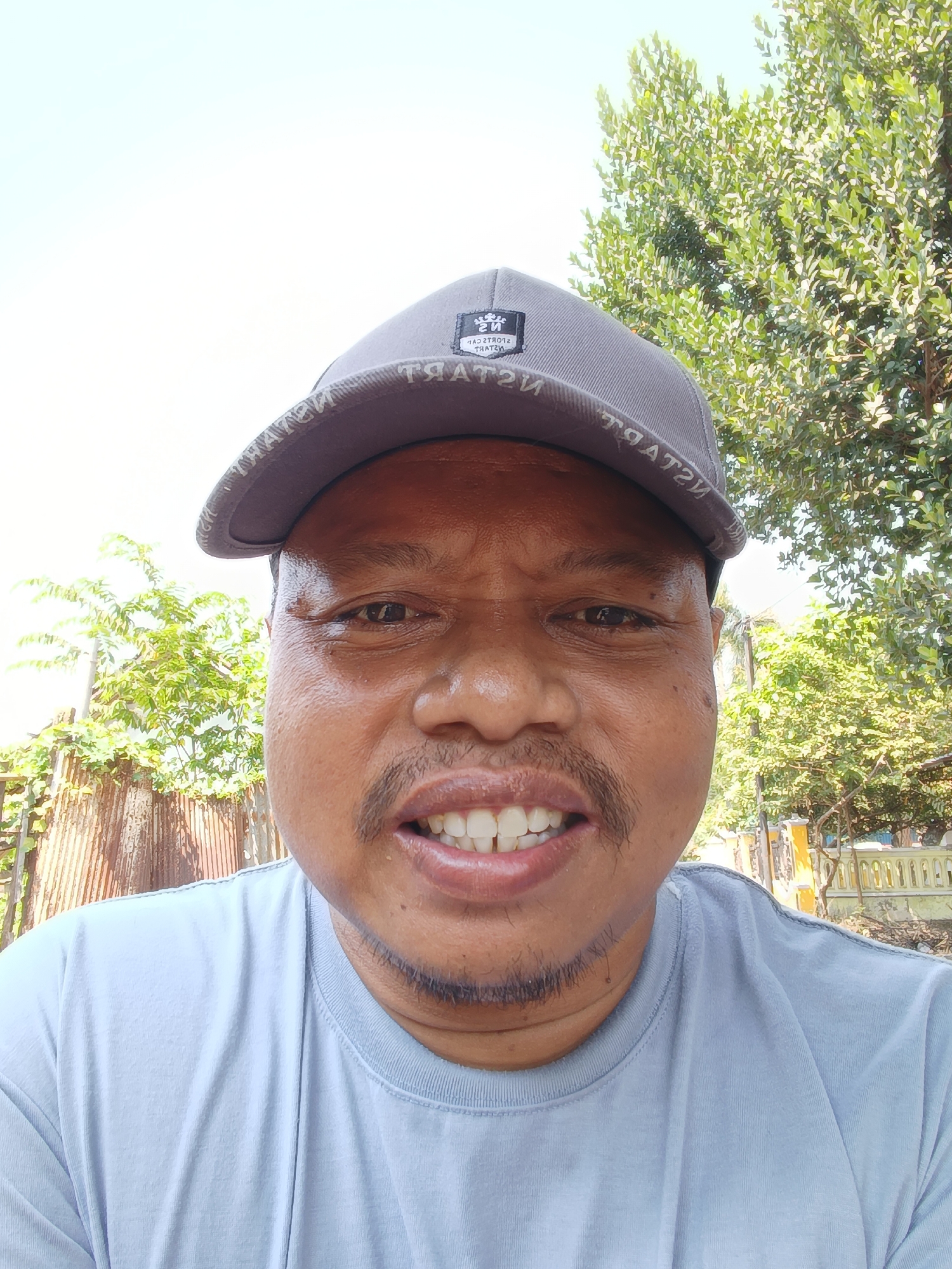 A close-up, waist-up, selfie-style photograph shows a man wearing a gray baseball cap and a light blue t-shirt. He has a light brown complexion, a mustache, and a small patch of stubble on his chin. His eyes are brown, and he has an open-mouthed smile, revealing his white teeth. The background features a bright sky, lush green trees, and some rustic fencing. The image appears to have been taken outdoors during the daytime, likely in a rural or suburban setting in Kandangjati Wetan, Indonesia.
