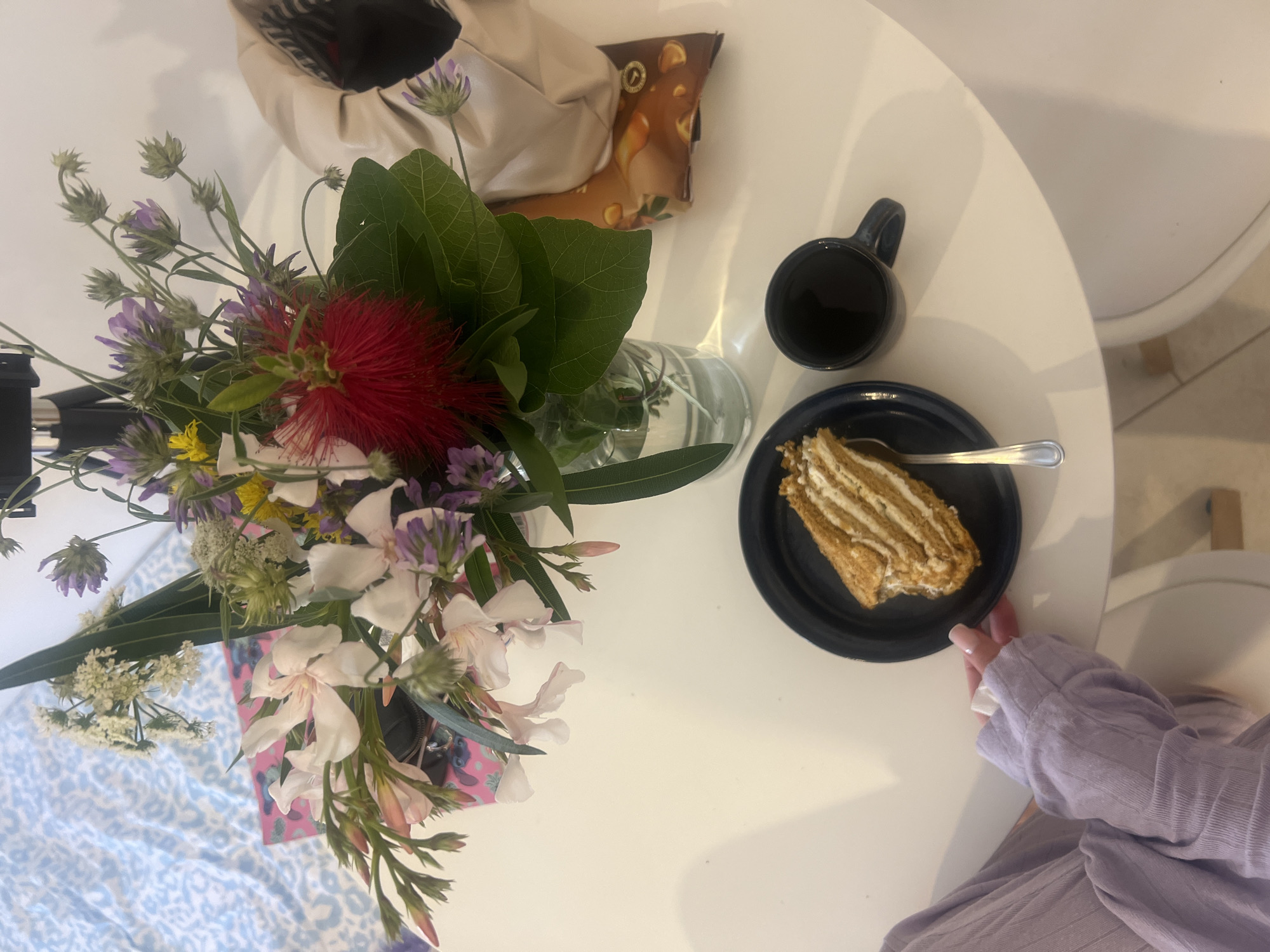 The image depicts an aerial view of a white round table. On the table, there's a bouquet of flowers in a vase, a cup of dark liquid, a slice of cake on a dark plate with a spoon, and a partially visible bag. A person wearing a light purple shirt is seated at the table. The location could potentially be a cafe or restaurant in the city of Budva, Montenegro, though specific architectural or geographic details confirming the location are absent.