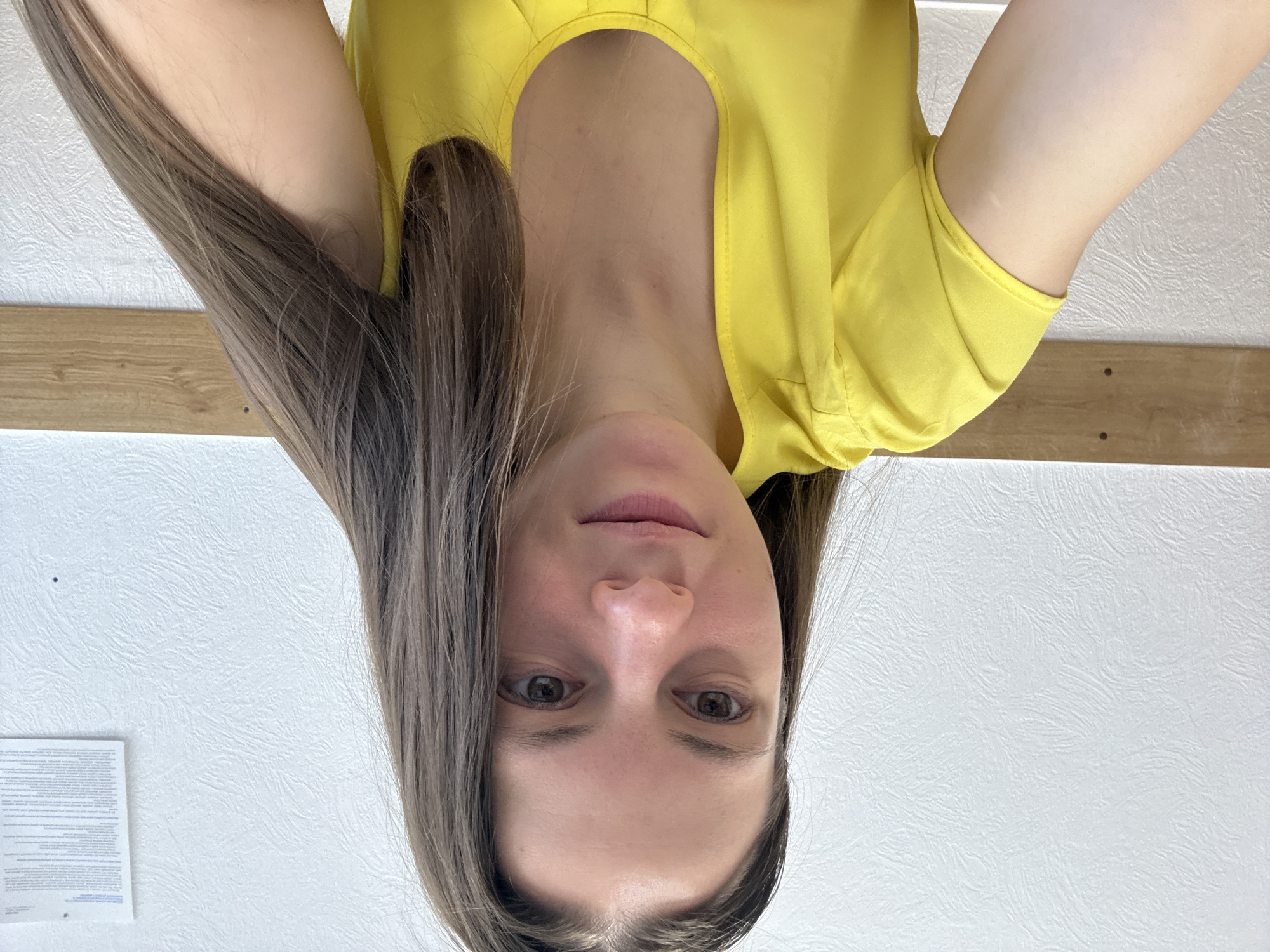 A woman with long, straight, brown hair is wearing a yellow top with a rounded neckline. The camera angle is from above, looking down at her face. The background consists of a textured white wall and a strip of wood trim. A document is affixed to the wall. This image was taken in the city of Chaikovsky, Russia.