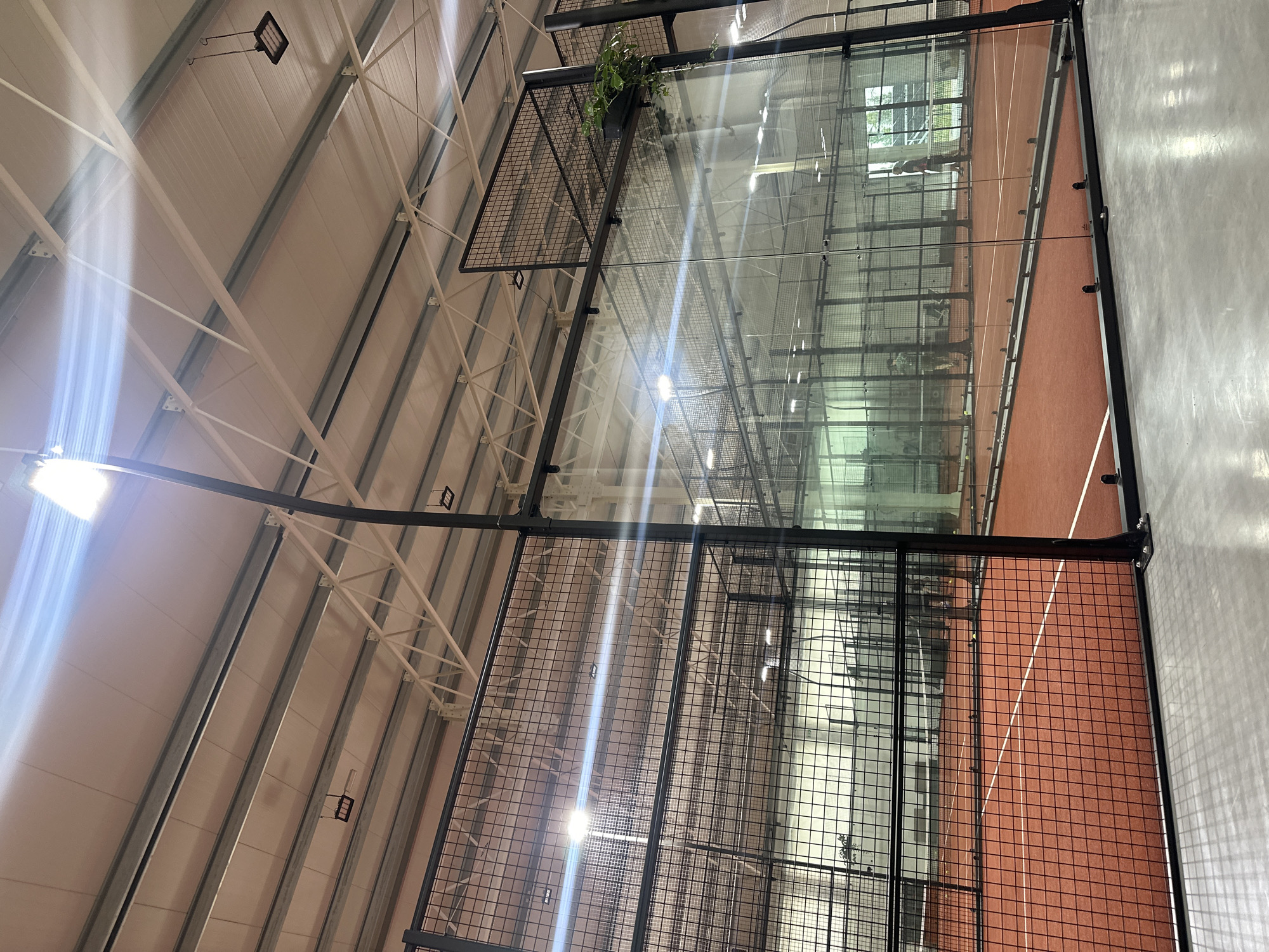 The image displays an indoor padel tennis court, featuring a terracotta-colored playing surface marked with distinct white lines. The court is enclosed by transparent glass walls and robust black metal mesh fencing, typical for this sport. Above, the ceiling of the facility showcases light-colored industrial paneling, supported by a network of white and grey exposed structural beams and girders. Numerous black LED panel lights are suspended from the ceiling, providing strong illumination to the court, with visible light flares originating from some of the active fixtures. A small green plant is placed in a black rectangular planter box on top of one of the mesh fences, adding a touch of greenery to the industrial environment. Faint reflections of individuals are discernible on the glass walls; one reflection shows a figure that appears to be holding a racket, suggesting the court is actively being used or is ready for a match. The overall scene depicts a modern and well-lit indoor sports setting. No visible text is present in the image.