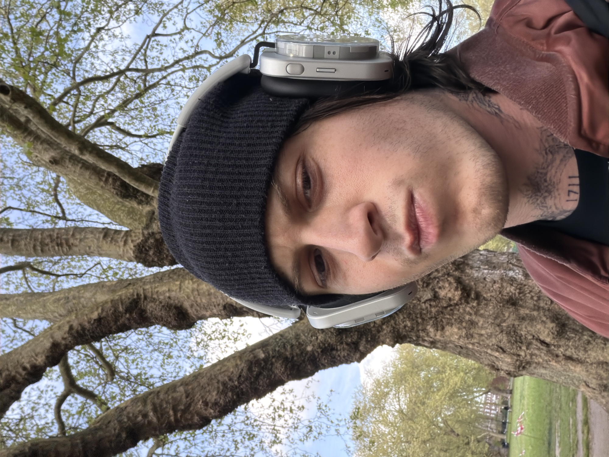 A self-portrait-style photograph captures a young adult male in a park setting, with the image rotated 90 degrees clockwise. The subject has fair skin, dark hair, and light facial stubble, looking directly at the camera with a neutral expression. He is wearing a dark grey knit beanie, atop which rests a pair of silver over-ear headphones featuring transparent earcups. He also wears a reddish-brown jacket. A prominent tattoo, including the numbers "1711," is visible on the right side of his neck.
The background is dominated by large, mature trees with thick, textured trunks and branches, adorned with sparse, light green spring leaves. A bright blue sky with scattered white clouds is visible through the tree canopy, suggesting a clear day. The perspective is from below, looking upwards towards the subject and the trees. Green grass and distant park elements, such as a path or bench, are faintly visible, consistent with an outdoor park environment in London, United Kingdom. The image appears to have been taken during daylight hours.
