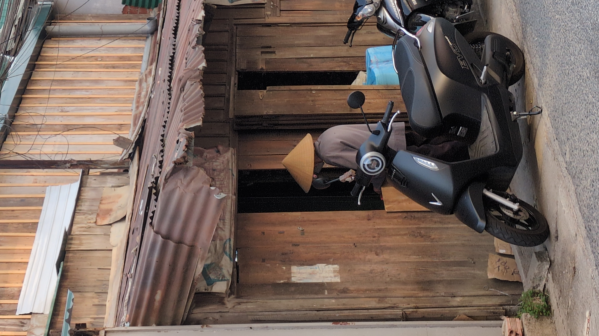 This image shows a person wearing a Vietnamese conical hat, standing in the doorway of a dilapidated wooden building next to a black VinFast Feliz S electric scooter, with another older scooter parked nearby. The building is constructed from wood and corrugated metal, showing signs of wear and decay. The location appears to be Da Lat, Vietnam, based on the traditional attire and architectural style. The person seems to be looking at or interacting with something behind the scooter. The scene is set on a narrow street or alley, with the building situated directly adjacent to the road. There is some trash or debris along the street.