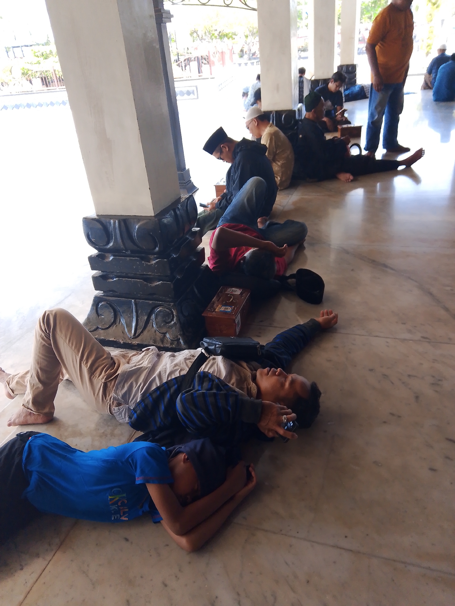 In Demak, Indonesia, an indoor scene depicts several individuals resting on a marble floor. In the foreground, two young men lie down. The one on the left is partially obscured, wearing a blue t-shirt and black pants, with his head resting on his arms. The man next to him, wearing a striped long-sleeved shirt and brown pants, is lying on his back, his head turned to the side, holding a dark object, possibly a phone, in his right hand. Further back, a man in tan pants and a light-colored shirt is also lying on the floor. A group of men are seated or lying down in the midground. One man, wearing a black skullcap and dark clothing, is seated and looking at his phone. Another man in a black skullcap and dark clothing is seated with his legs extended. A man in a yellow shirt stands in the background, facing away from the camera, while another man in blue sits with his back to the viewer. The environment appears to be an open area with columns and natural light filtering in. The overall impression is one of people relaxing or waiting in a public indoor space.