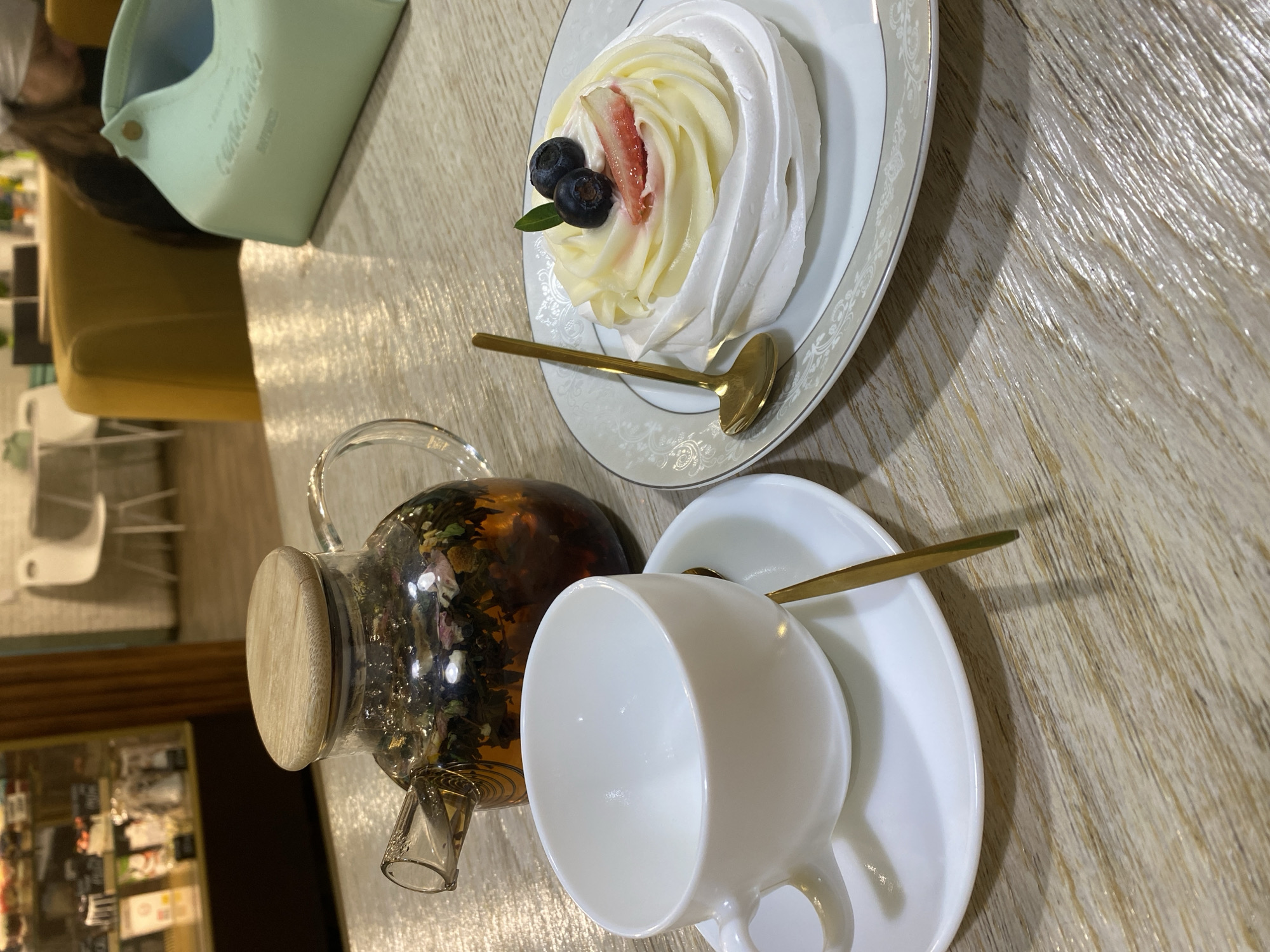 A static, eye-level image depicts a table setup within a commercial establishment, specifically a cafe or restaurant interior. The precise location within Izhevsk, Russia, is not ascertainable from the visual information provided.
On a light wood-grain table surface, a clear glass teapot with a circular wooden lid is present on the left, containing brewed herbal tea with visible dark leaves. Adjacent to the teapot, a white ceramic teacup sits empty on a matching saucer, accompanied by a small golden spoon. To the right, a white dessert plate with an ornate grey and silver border displays a meringue-based dessert. This dessert features a white meringue base, topped with piped white or cream-colored frosting, two dark blueberries, one sliced strawberry, and a small green leaf garnish. A second small golden spoon rests on this dessert plate.
In the background, a display case with various pastries is partially visible on the left. On the right, a person with dark hair, wearing a light-colored head covering, is seated in a yellow upholstered chair, facing away from the camera. A light mint-green, structured bag or container is positioned on the table to the right of the person. Further into the background, additional white chairs, tables, and a white brick-patterned wall indicate a larger seating area. No direct actions or interactions are occurring with the items on the table.