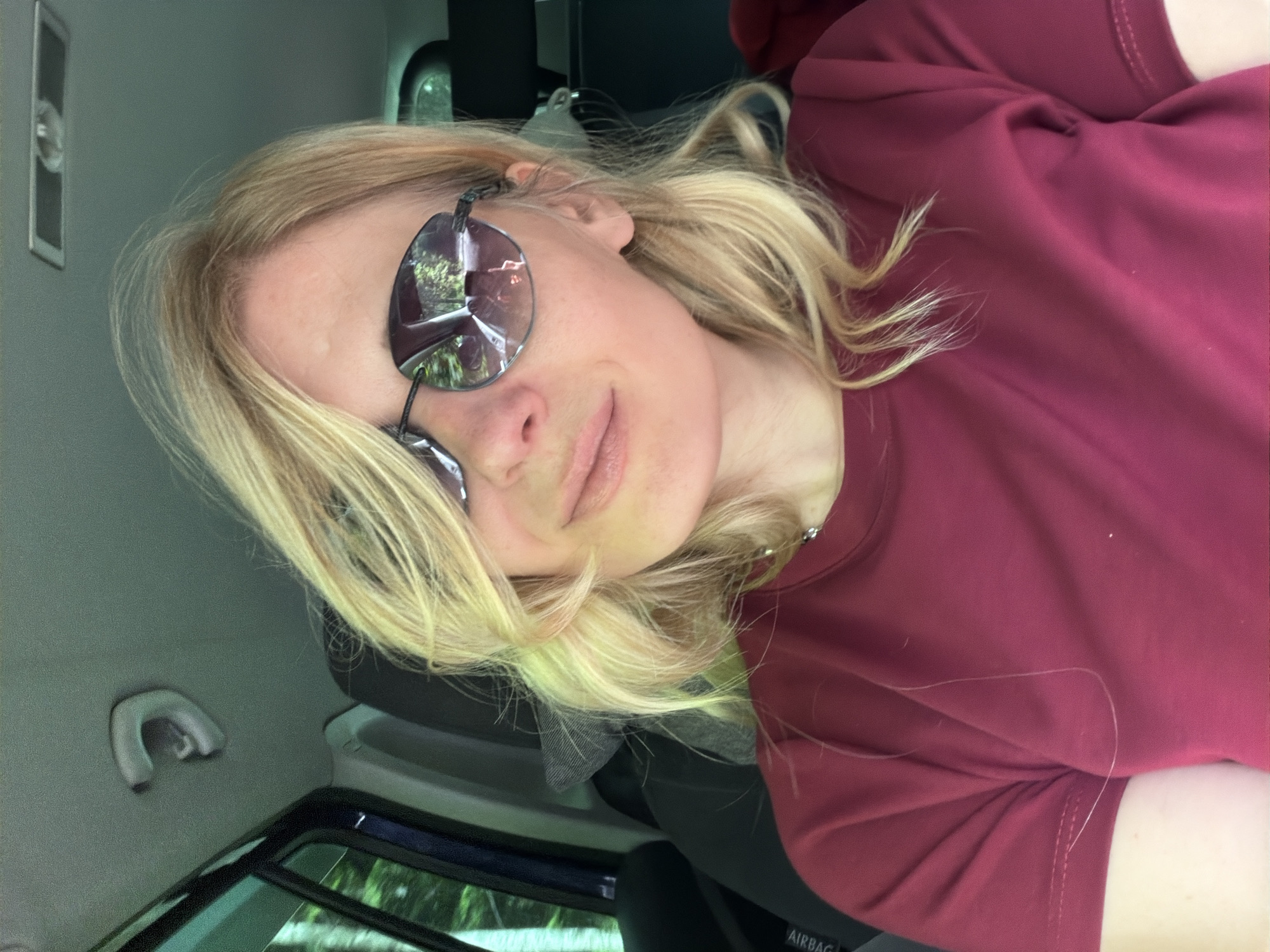 The image shows a blonde woman with sunglasses in the front passenger seat of a car. She is wearing a maroon t-shirt and has her hair down. The car is a dark color, and the interior appears to be gray. The woman is looking at the camera and smiling slightly. The background of the image is blurred, but it appears to be a wooded area. The image was taken in Верхне-Тебердинское сельское поселение, Russia.