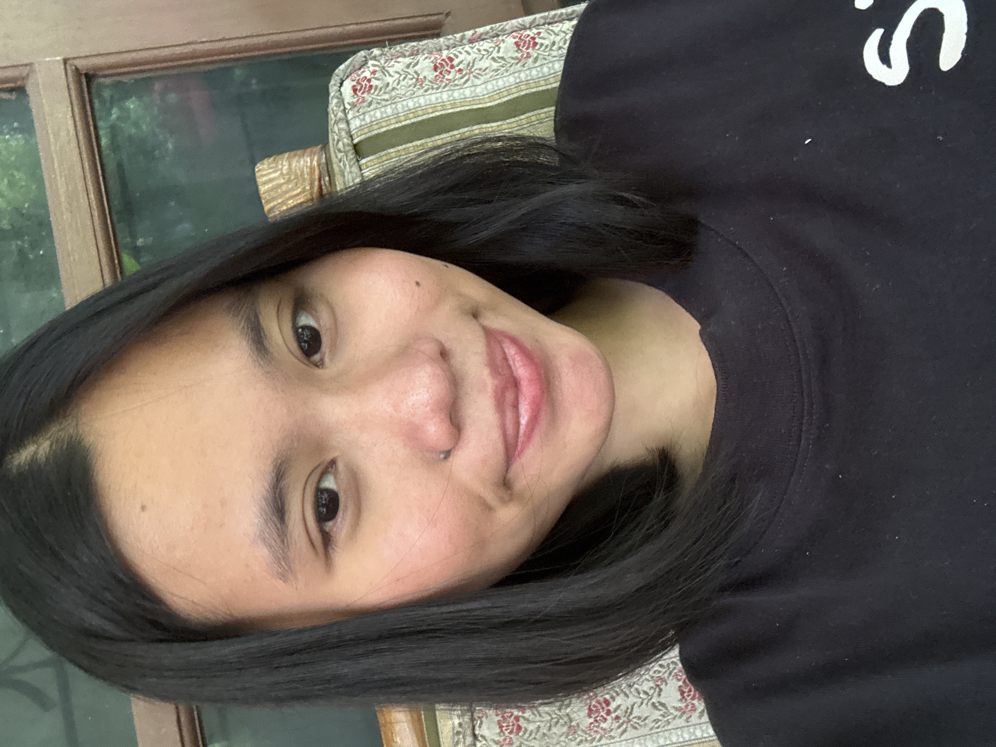 A young woman with medium-length black hair and a nose piercing is pictured in a close-up, front-facing portrait. She is wearing a black t-shirt. The background suggests an indoor setting with natural light, possibly a room with a window and furniture. The woman's facial expression is neutral to slightly smiling. The overall lighting is soft and appears to be from a natural source.