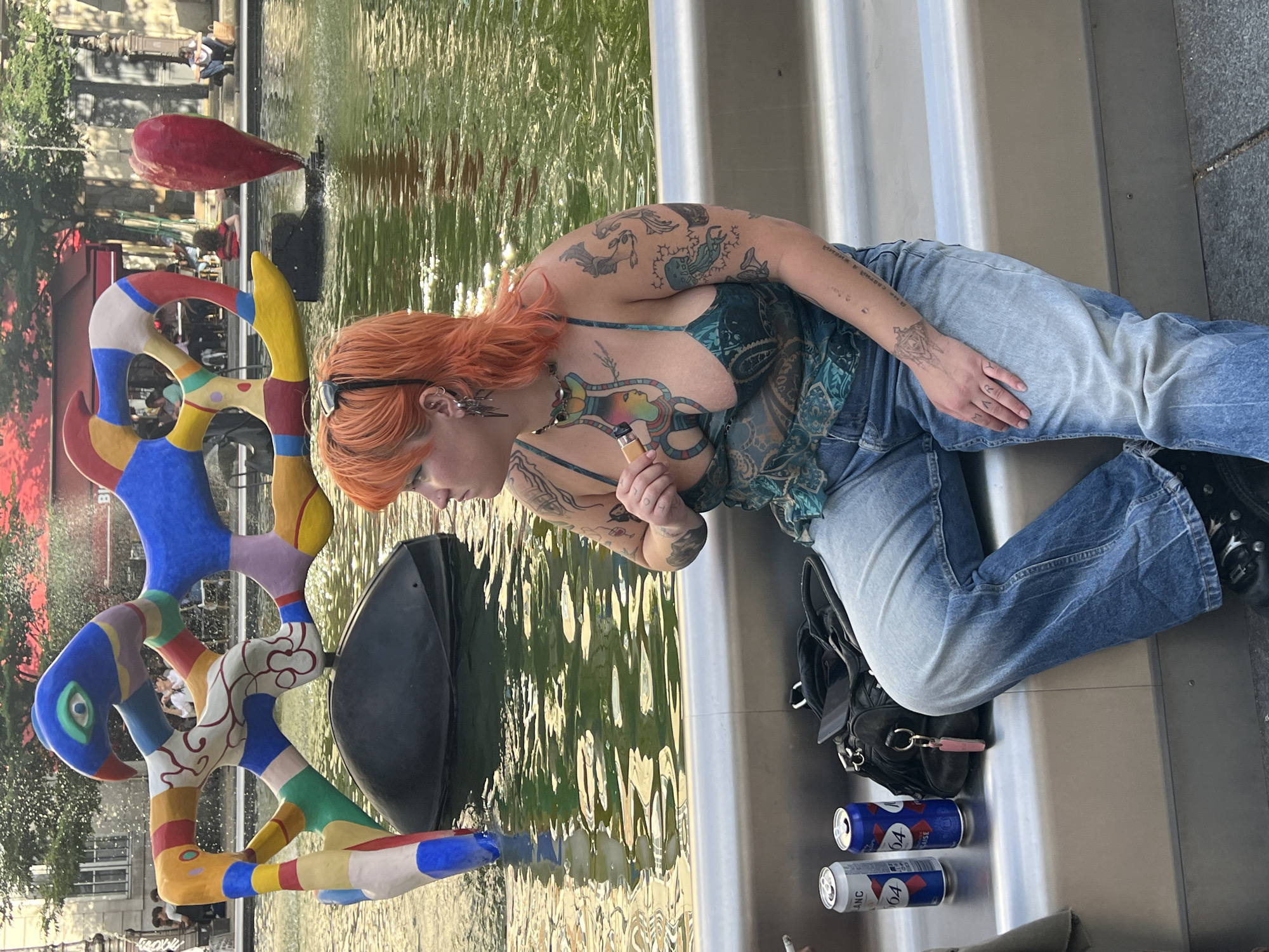 This is a photograph of a woman with orange hair, tattoos, and piercings sitting on a metal bench in front of a fountain. She is wearing a blue and green patterned tank top and blue jeans. Next to her are two cans of beer, a black bag, and a pink keychain. In the background, there is a colorful abstract sculpture in the fountain and buildings. The fountain is located in the Place de la Fontaine des Innocents in Paris, France.