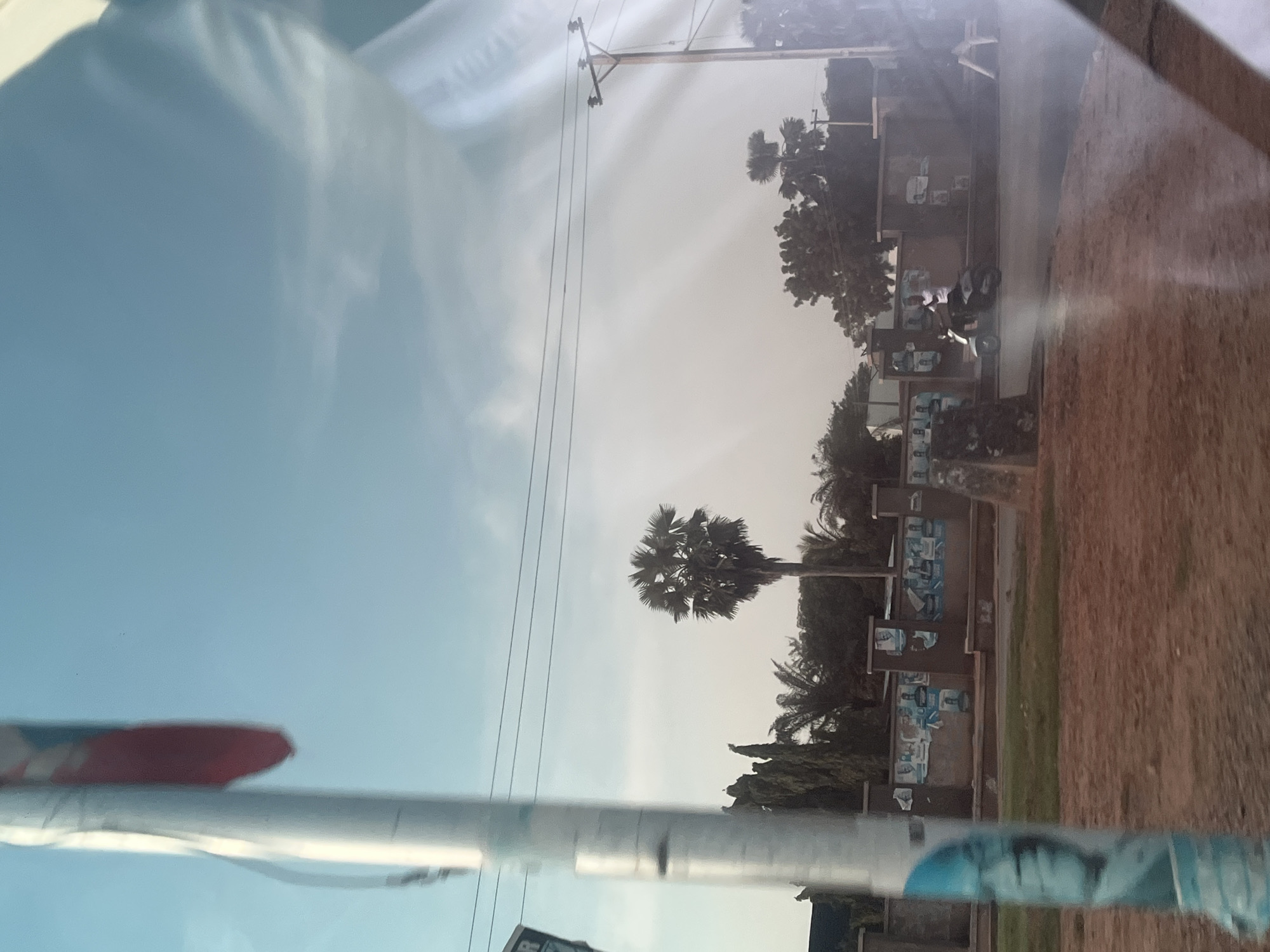 The image depicts a street scene in Jalingo, Nigeria. A prominent, out-of-focus flagpole with a tattered red and white flag is in the foreground on the left. Beyond the flagpole, a paved road runs horizontally. A person is riding a scooter on the road. To the right of the road, there is a wall adorned with posters, some featuring portraits of individuals. Behind the wall, trees, including a distinctive palm tree, are visible against a bright, hazy sky with light clouds. Electrical wires and a utility pole with insulators are also present in the sky. The ground in the foreground is a reddish-brown, appearing to be dirt or gravel.