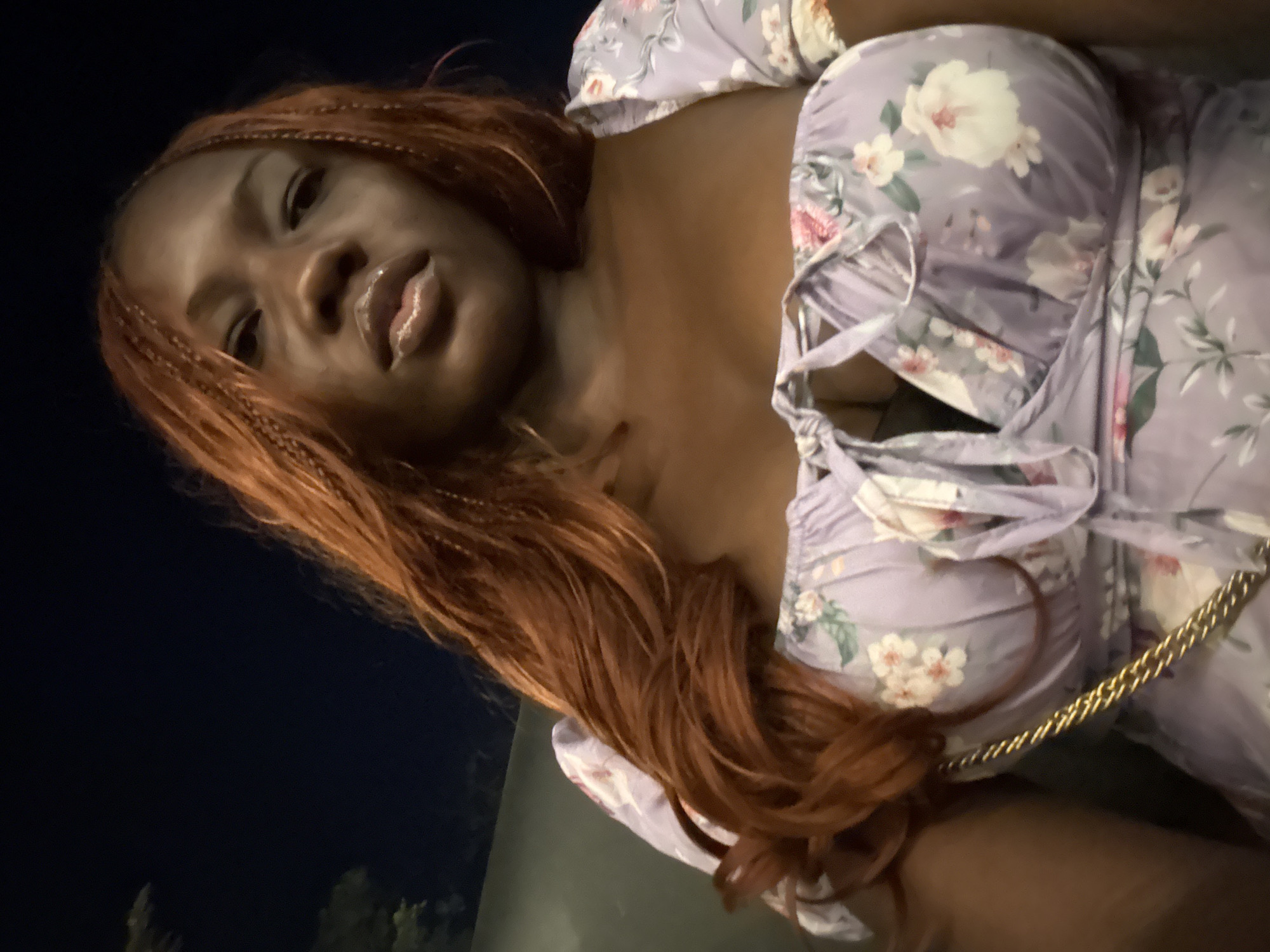 A young Black woman is prominently featured in a close-up, slightly tilted portrait, possibly a selfie, set against a dark, nighttime background in Abuja, Nigeria. She looks directly at the camera with a neutral expression, her lips appearing glossy. Her long, reddish-brown hair, styled in braids or twists, cascades over her shoulders. She is wearing a light purple or lilac dress adorned with a white, pink, and green floral pattern. The dress features a gathered or ruched bodice with visible tie details on the front. A gold chain, likely from a handbag or other accessory, is visible across her chest. The lighting suggests an artificial source illuminating the subject, contrasting with the dark background which indicates it is nighttime. Blurry green elements at the bottom of the frame might be foliage or trees in the outdoor setting. No text is visible in the image.