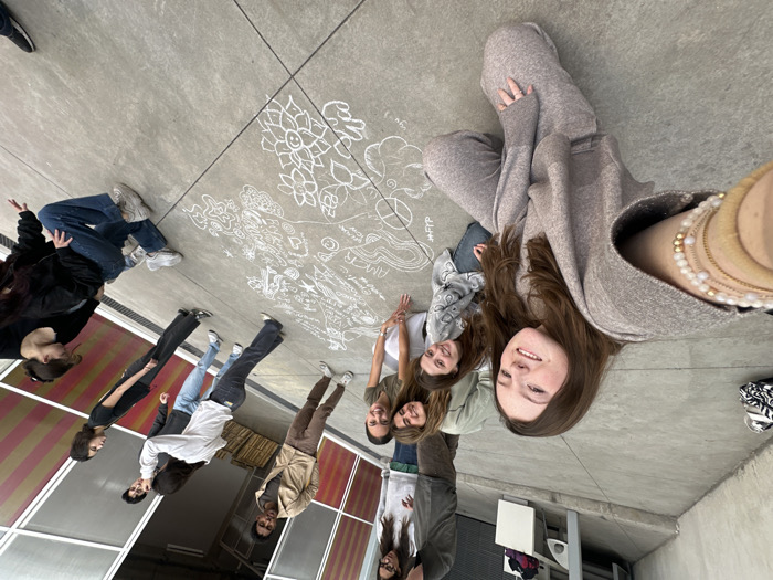 A group of approximately twelve young individuals, primarily women, are depicted in an outdoor concrete courtyard or corridor in the City of Unknown, Unknown. The central focus is a large, intricate chalk drawing on the concrete pavement, featuring various patterns, symbols including a peace sign, hearts, flowers, and text such as "AMOR" and "ffpp." Several individuals are kneeling or crouching around the drawing, suggesting active engagement in its creation or observation.
In the foreground, a young female with long brown hair, wearing a light brown knit two-piece outfit (wide-sleeved top and matching pants), kneels and smiles directly into the camera, taking a selfie. Her left wrist displays multiple bracelets, including gold-toned and white beaded styles. To her right, another young female kneels, also smiling towards the camera, dressed in a grey patterned long-sleeved top and blue jeans.
Behind them, other group members are positioned around the chalk art. These include: a young woman in a light green top and light pants; a young woman in a black top and blue jeans, kneeling on the far right; and several individuals standing or interacting in the mid-ground. Among the standing figures are two males: one in a light brown jacket and dark pants, and another in a dark grey top and light blue jeans. Two females are also standing, one in a white long-sleeved top and dark jeans, and another in a black top and wide-leg black pants.
The background features a building facade with a modern design, characterized by a grid of translucent or frosted panels, some exhibiting a reddish-brown hue and others appearing clear or white. To the upper left, an industrial-style grey wall is visible with a white bench or ledge. The ground is a uniform grey concrete surface. A black and white patterned bag is on the ground near the left edge of the frame. The scene is evenly lit, indicating daytime conditions.