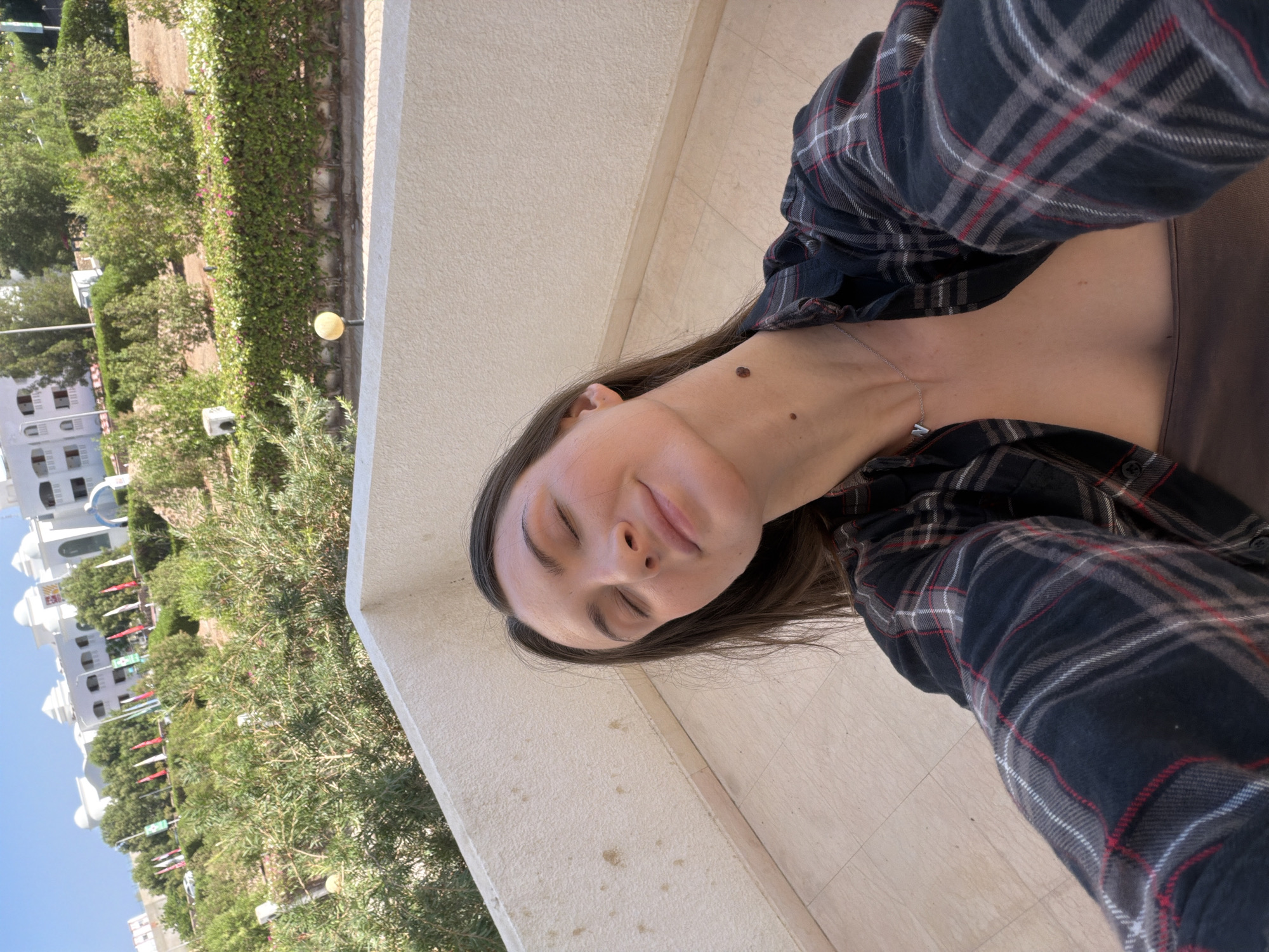 A young woman with brown hair is taking a selfie outdoors. She is wearing a plaid shirt and a necklace. Her eyes are closed and she appears to be smiling. She is on a balcony overlooking a green landscape with buildings and flags in the background. The location is Sharm El Sheikh, Egypt.