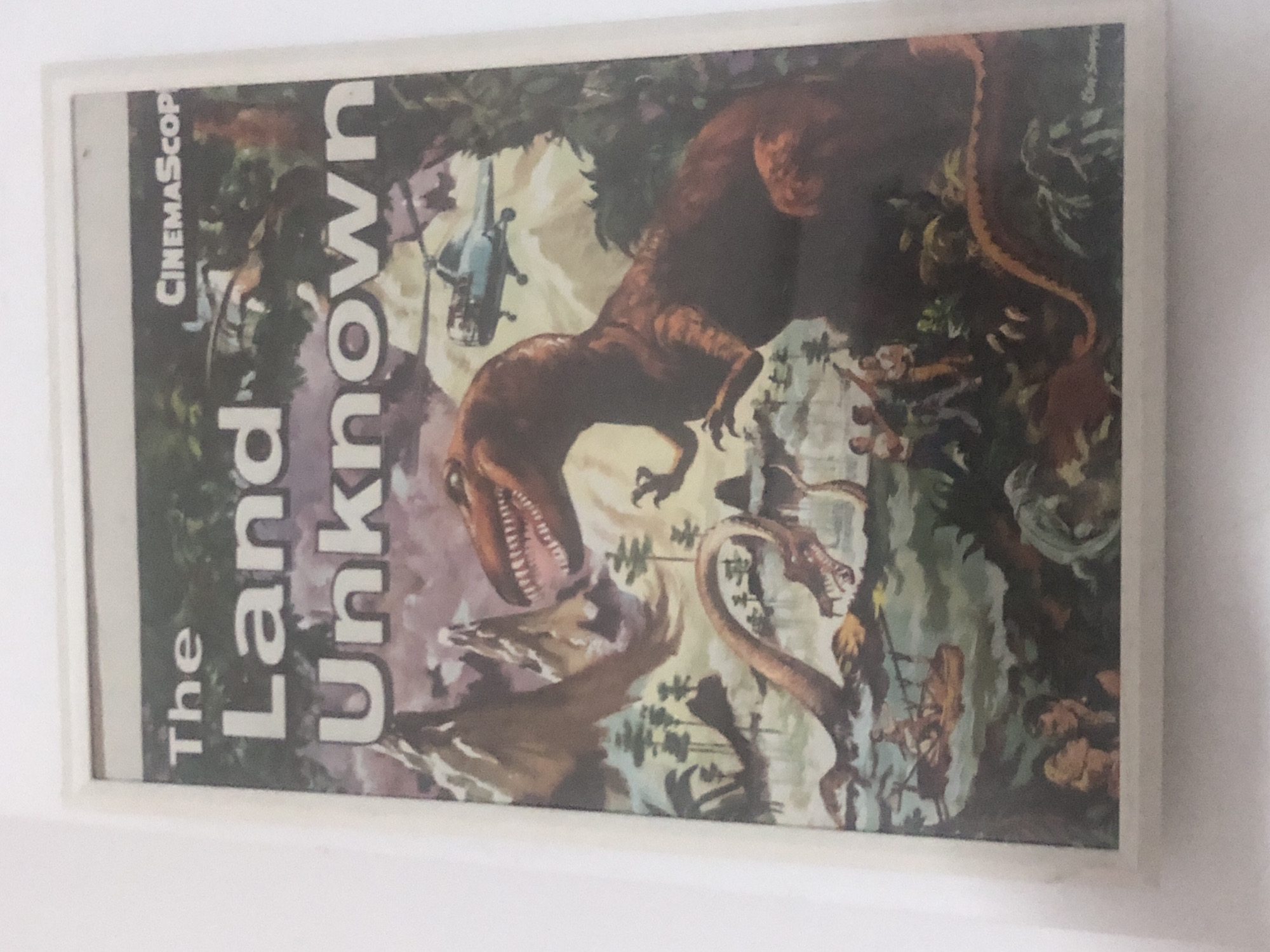 The image is a painting of a prehistoric landscape with several dinosaurs. A large red Tyrannosaurus Rex stands in the foreground, facing right. To its left, a long-necked Plesiosaur emerges from the water, with its head turned towards the Tyrannosaurus. In the background, a helicopter flies over a mountainous, jungle-covered terrain. A group of people are visible in the lower right of the image, observing the scene. The title of the painting, "The Land Unknown," is written in large white letters at the top of the image, with "CinemaScope" written in smaller letters to the right of the title. The overall style of the painting is vibrant and detailed, with a sense of action and adventure.