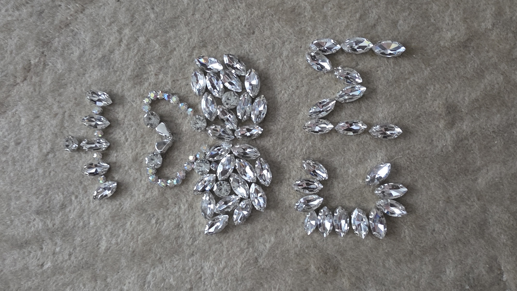 The image displays an artistic arrangement of numerous clear, faceted rhinestones and crystals on a light brown, soft-textured surface, possibly carpet or felt. The crystals are predominantly marquise-shaped, with a few small round ones interspersed.
On the far left, a vertical line of five marquise crystals, augmented by two small round crystals, distinctly forms the number '1'. To its right, a larger, more intricate cluster of marquise and round crystals is present, partially outlined by a string of iridescent beads. This central formation appears to be a stylized '0' or an abstract decorative element. Further to the right, marquise crystals are arranged to clearly form the letter 'S'. Below the 'S', a semicircular or C-shaped arrangement of marquise crystals completes the composition, possibly serving as an additional decorative element or part of an incomplete symbol.
The crystals reflect light, creating a sparkling effect. The scene is static, showcasing a meticulously placed arrangement, implying a creative or decorative activity. No people are visible, and the setting appears to be indoors. The photo was taken in Blagoevgrad, Bulgaria.