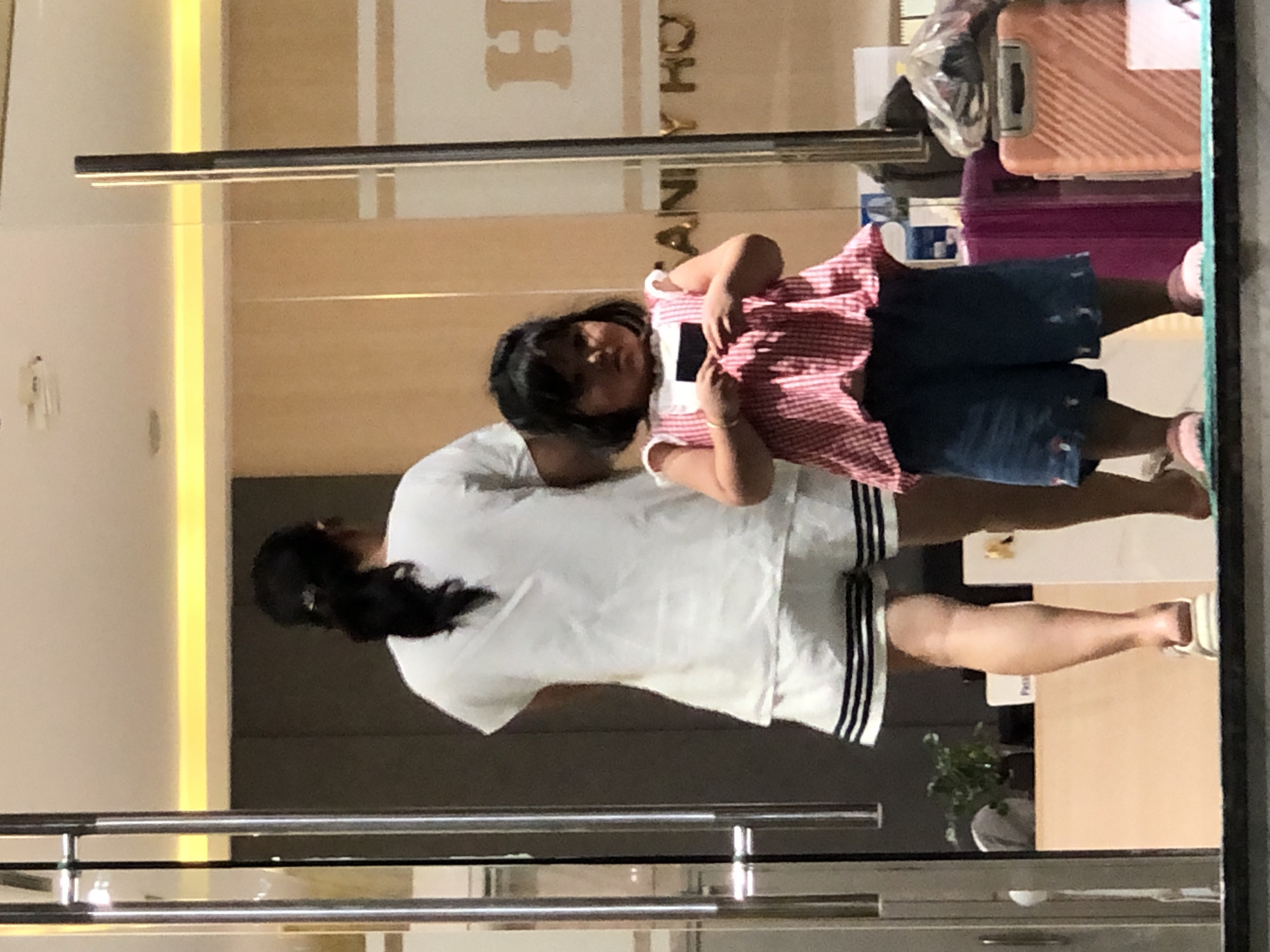 The image depicts two individuals, an adult and a child, standing inside a building that appears to be a hotel lobby in Đà Nẵng, Vietnam. The adult, seen from behind, has dark hair pulled back into a ponytail and is wearing a white t-shirt and white shorts with black stripes. The child, facing the camera, has dark hair and is wearing a red and white checkered top and blue shorts. The child is holding a dark object, possibly a credit card or ID. The background shows elements of a hotel, including glass doors, wooden paneling, and signage that reads "H AN Y HO". Luggage is visible to the right of the child, including a pink suitcase and a peach-colored suitcase. The lighting suggests an indoor environment, possibly during the day or evening.