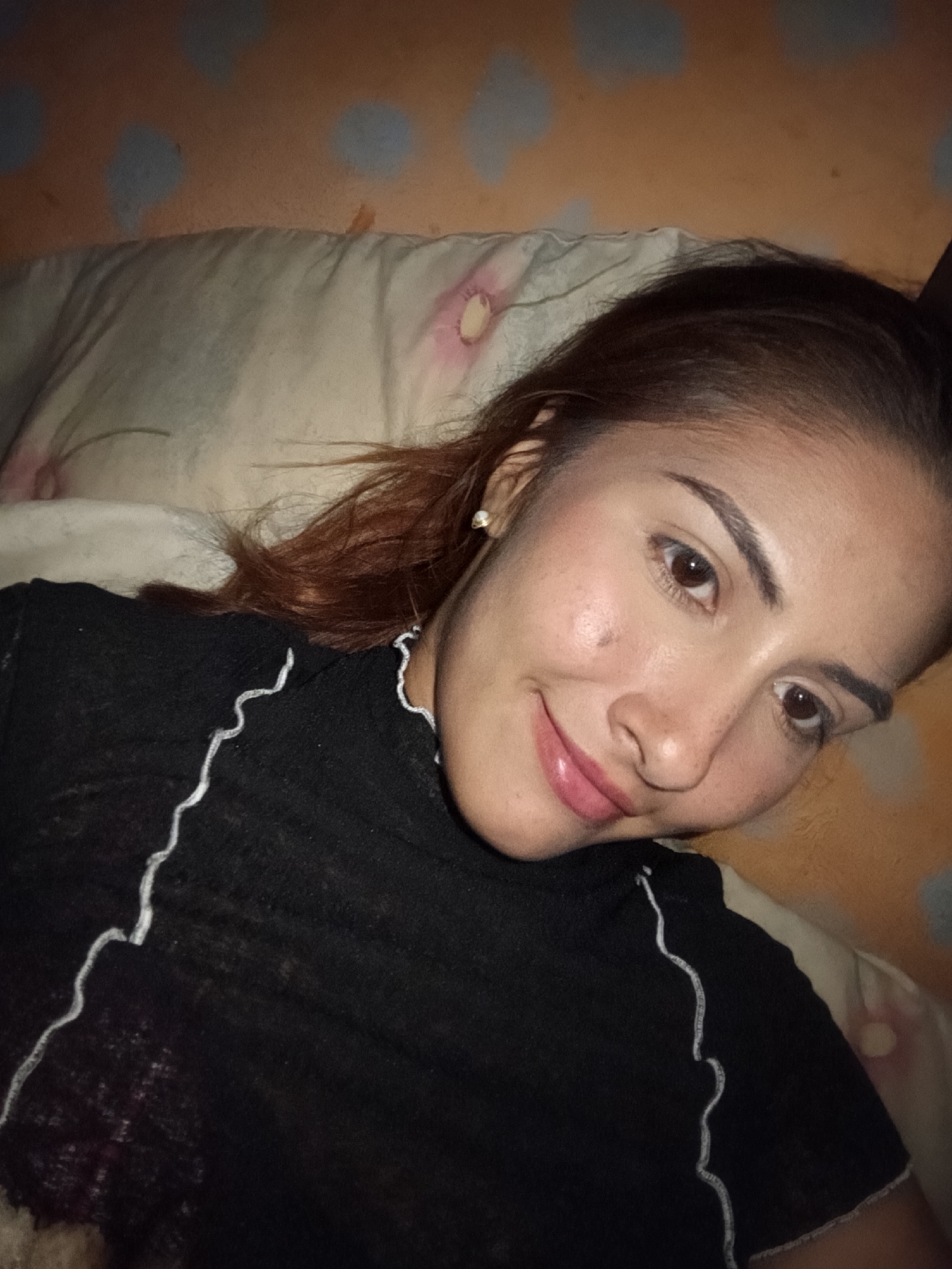 A young adult female with a fair to medium complexion, brown eyes, and brown hair with subtle reddish highlights is depicted in a close-up, top-down perspective. She is lying down, looking towards the camera with a relaxed, gentle smile. She wears a black long-sleeved top characterized by prominent white wavy stitching along the neckline and down the side seams. A small, spherical pearl earring adorns her left ear. A singular dark mole or freckle is visible on her upper left cheek. Her hair is spread upon a light-colored pillow, which shows faint, light pink or purple patterns. Behind the pillow, an orange or tan surface is visible, patterned with several diffused, blurred blue circular shapes. The specific location in the city of Juan Griego, Venezuela, cannot be determined from the visual information provided in the image.