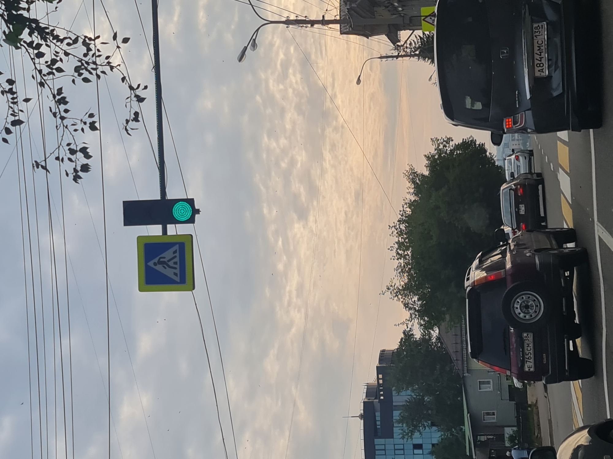 The image depicts a street scene in Irkutsk, Russia, on a cloudy day. A traffic light with a green illuminated circle is visible, alongside a pedestrian crossing sign. Several cars are on the road, including a maroon SUV with a spare tire mounted on its rear and a dark minivan with its license plate clearly readable as A844MC 138. In the background, there are buildings, trees, and utility poles. The sky is overcast with some lighter patches, suggesting late afternoon or early morning light. The perspective is from inside a vehicle, looking forward.