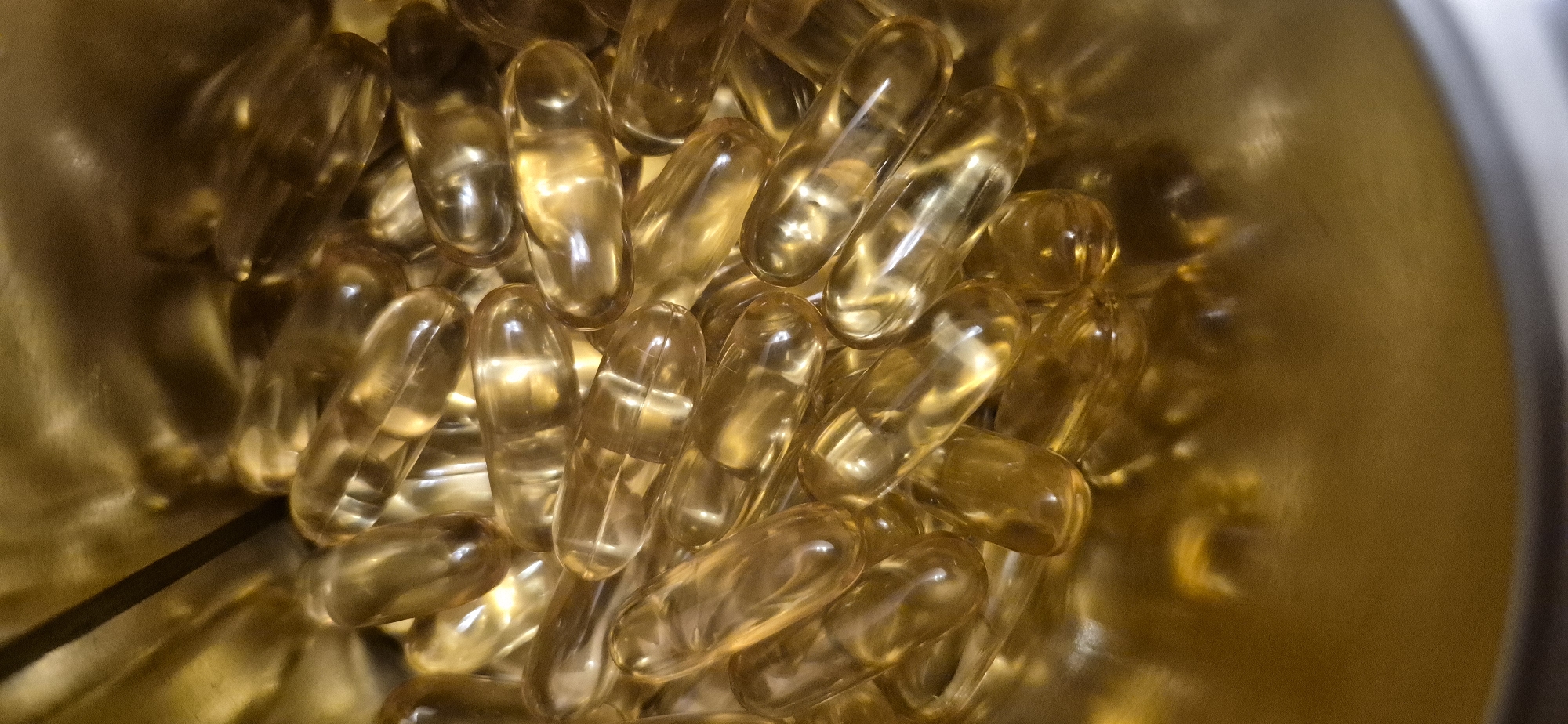 This is a close-up, overhead shot of a pile of softgel capsules, likely supplements such as fish oil or vitamin E, filling the bottom of a metallic, golden container. The capsules are translucent, oval-shaped, and appear to be filled with a yellowish liquid. The lighting creates reflections on the capsules and the container, giving the image a warm, golden hue. There are no people or any indication of a specific location. Therefore, it is not possible to provide information about the city of Baden-Baden, Germany, or any forensic details of a scene.