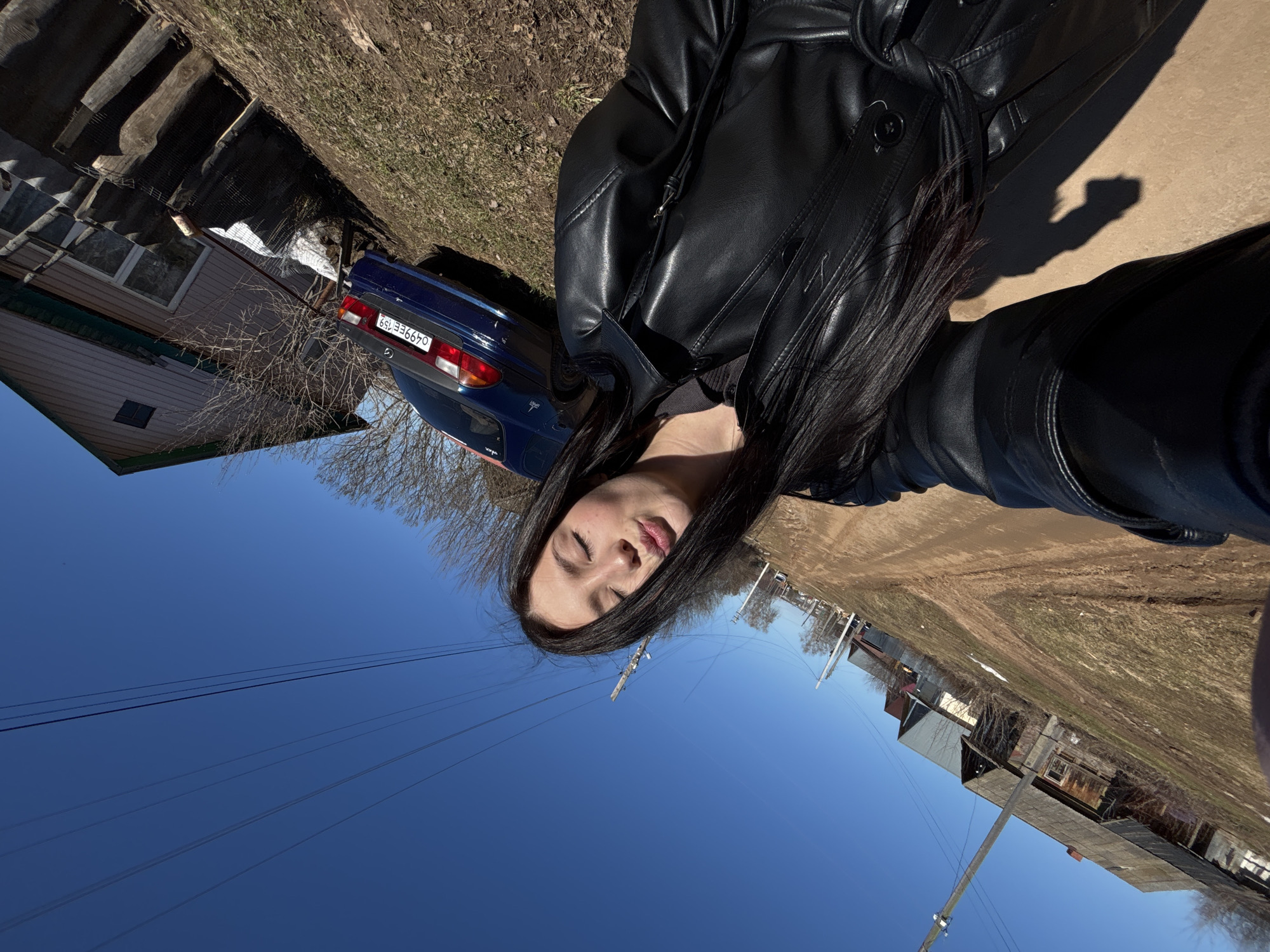The image depicts a woman with long, dark hair and a black leather jacket in an outdoor setting in Фоки, Russia. She is positioned in the foreground, and behind her is a blue car parked near a building with a visible license plate that reads "O499EE 159". Power lines are overhead, set against a clear blue sky. The background includes residential buildings and a dirt road. The lighting indicates a sunny day, and a shadow is cast by the person taking the photo.