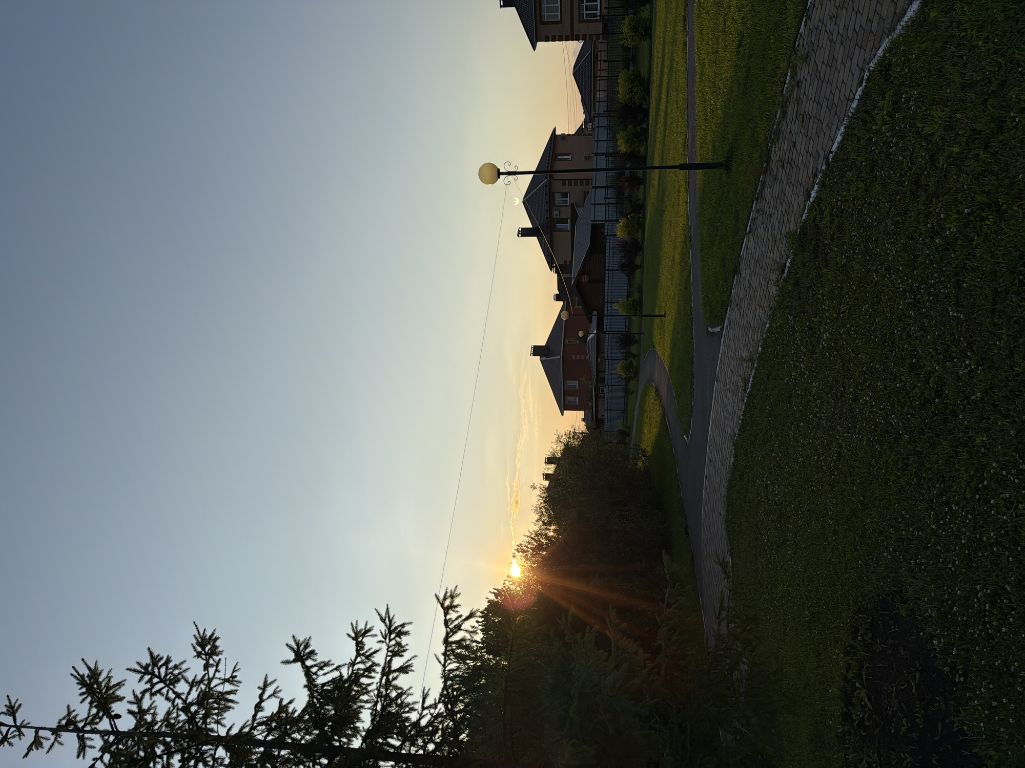 The image depicts a sunset scene in a suburban area, possibly a housing development, located in Shmidrovo, Russia. The sun is low on the horizon, casting warm, golden rays through trees and clouds. Silhouetted pine trees frame the foreground, adding a natural element to the scene. In the middle ground, a row of modern brick houses with dark roofs are visible, separated by fences and manicured lawns. A paved walkway curves through the green grass, and a lamppost stands near the houses. The sky is a soft gradient of blue and orange, indicating either early morning or late evening. The overall atmosphere is serene and tranquil, with a sense of peaceful domesticity. No text is visible in the image.
