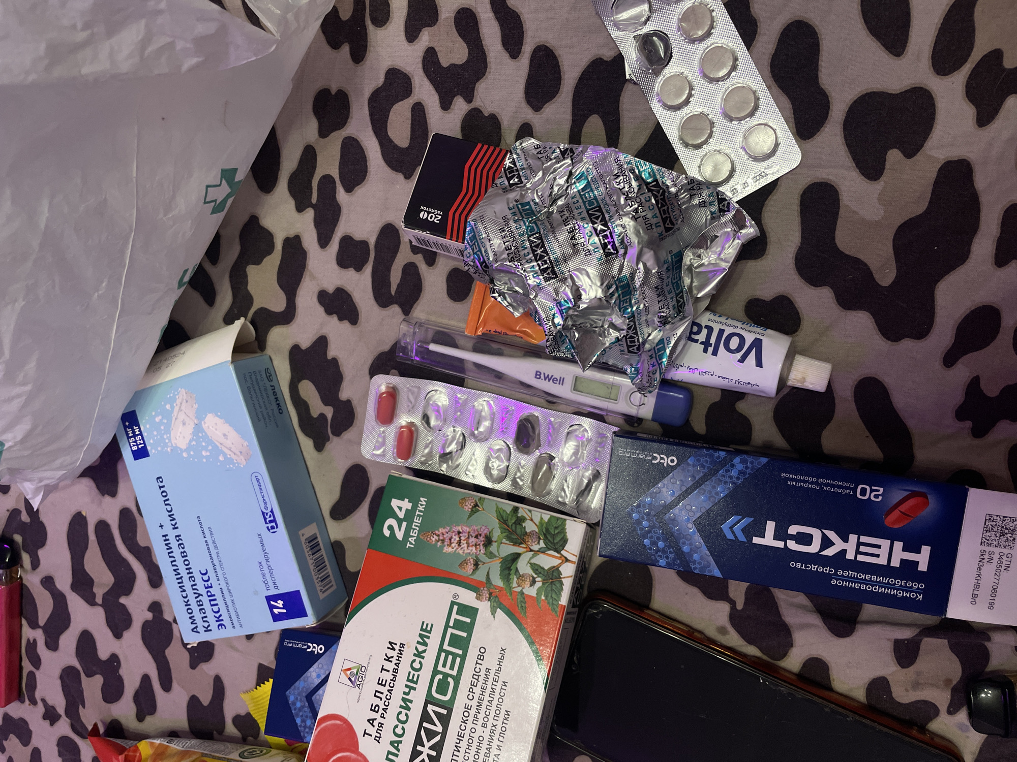 The image displays a collection of various medications and personal items scattered on a camouflage-patterned surface. Prominently featured are several boxes and blister packs of pills, including what appears to be an antibiotic box labeled "АМОКСИЦИЛЛИН + КЛАВУЛАНОВАЯ КИСЛОТА ЭКСПРЕСС" (Amoxicillin + Clavulanic Acid Express). Another prominent box is for "ЛАССИЧЕСКИЕ" (Lassicheskie) lozenges. There is also a box labeled "НЕКСТ" (Nekst), which contains 20 coated tablets. A digital thermometer, a tube labeled "Volta," and a smartphone are also visible among the medical supplies. The overall arrangement suggests a personal collection of medications. The image does not provide any information about the location in the city of Irinovka, Russia.