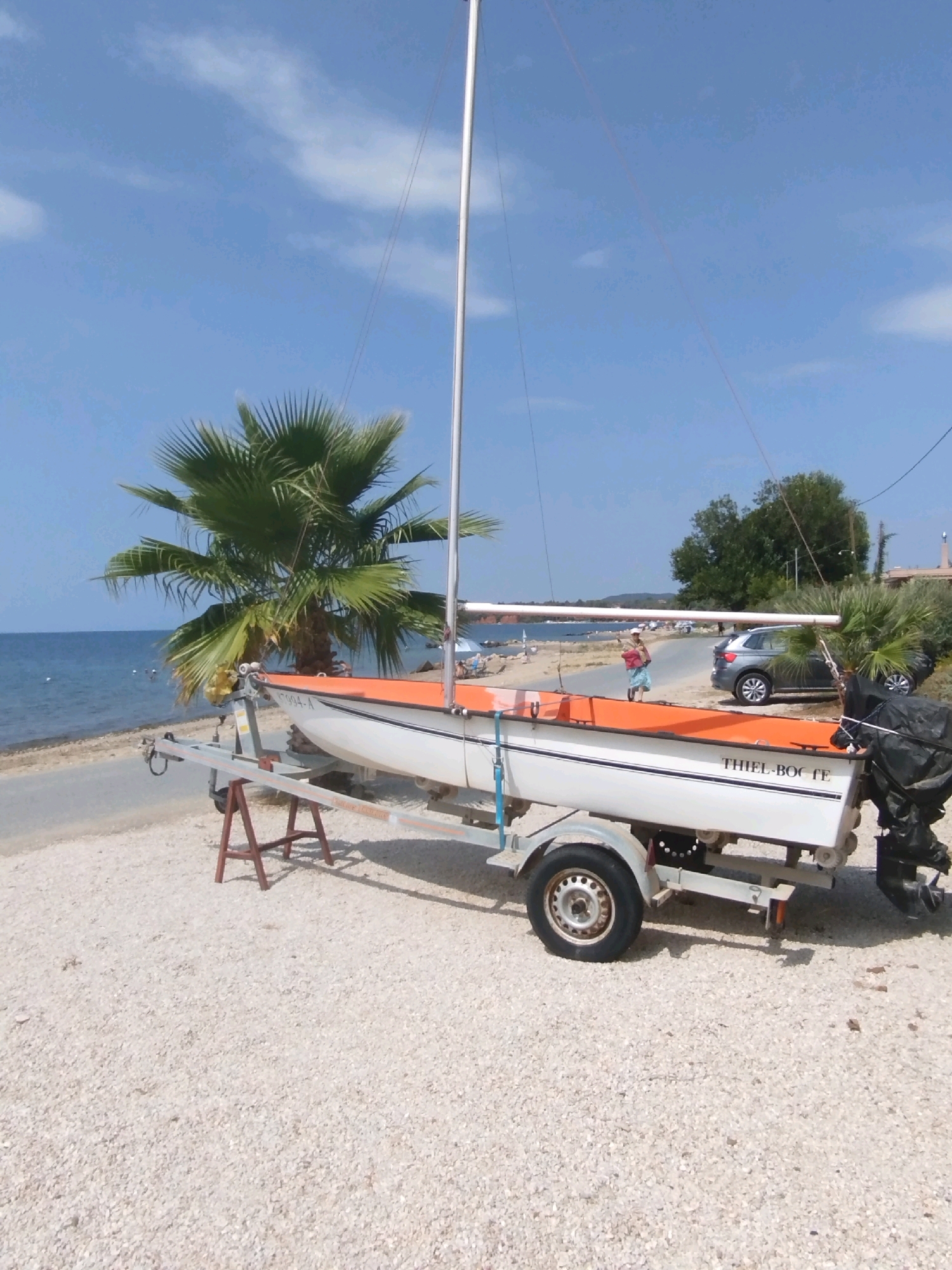A white sailboat with an orange trim is parked on a trailer on a gravel beach next to the sea. A palm tree is visible behind the boat. In the background, there is a road with cars and a few people. The sky is blue with some clouds. The boat has the inscription "THIEL-BOCFE" on its side. There is a black outboard motor attached to the stern of the boat. The trailer has two red A-frame supports underneath.