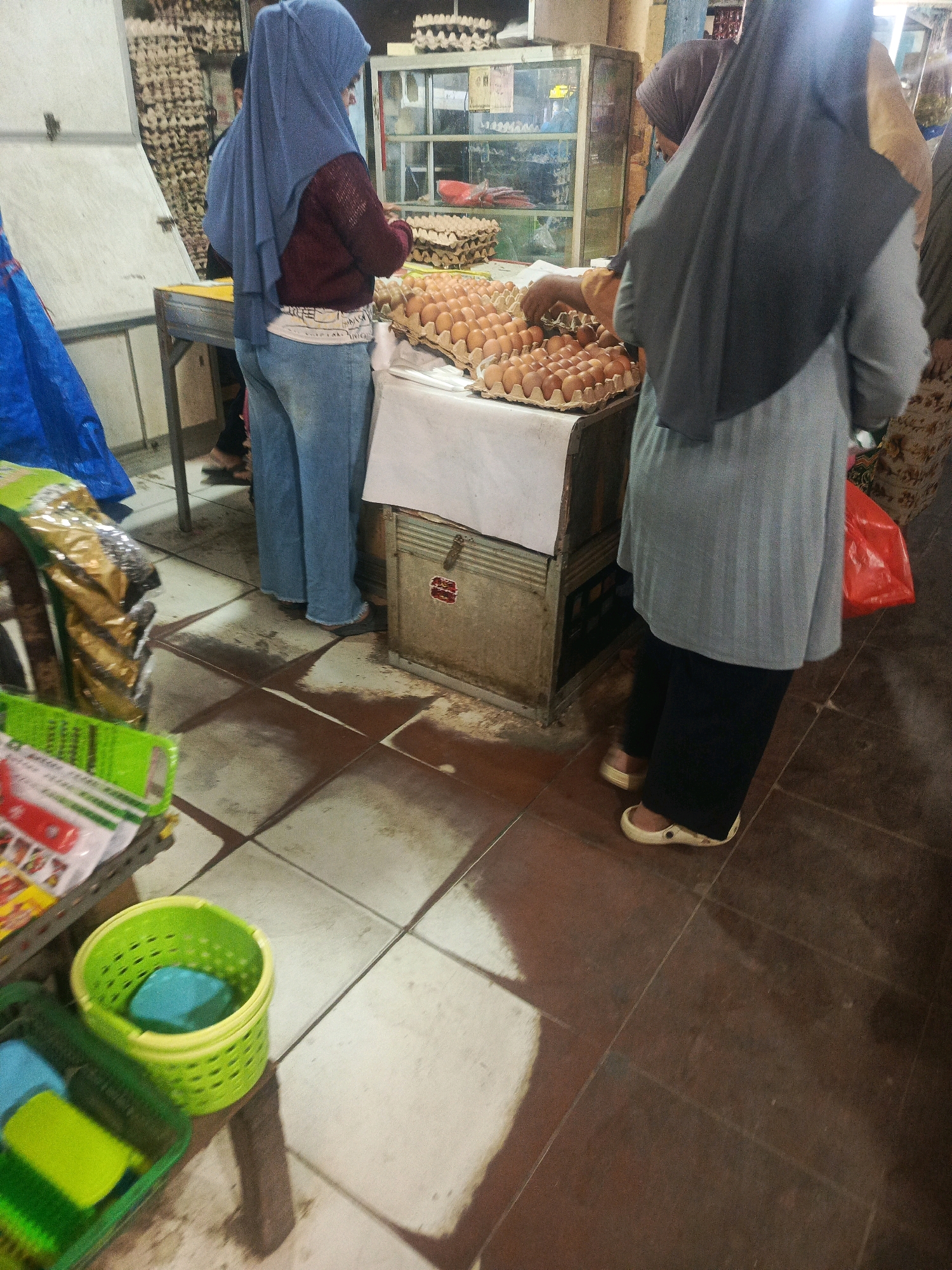 In Kendari, Indonesia, a market scene unfolds. Two individuals are visible. One, wearing a blue hijab and a maroon sweater over a white shirt, stands in front of a table piled with trays of eggs. They are also wearing blue jeans and dark footwear. The second individual, with a dark gray hijab and a light gray tunic over black pants and light-colored clogs, is seen from the back. They appear to be interacting with the eggs. In the background, a display cabinet with shelves is visible, and beyond it, racks of egg cartons are stacked high. A blue tarp is present on the left, and a red plastic bag hangs on the right. The floor is tiled, with some areas appearing damp or stained. In the foreground, green plastic baskets are arranged, one holding a blue object.