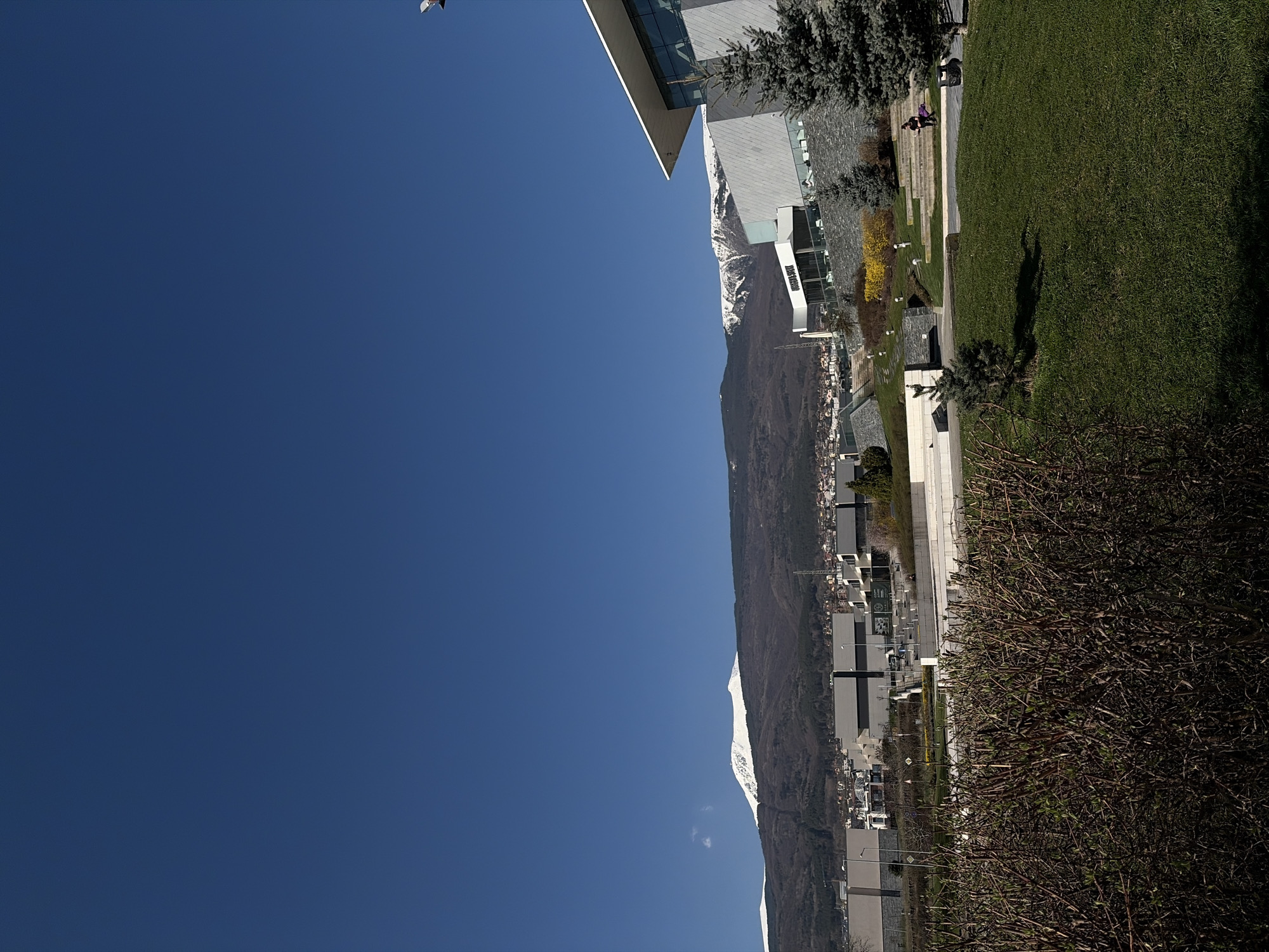 The image showcases a bright blue sky with a distant mountain range along the horizontal axis. Adjacent to the mountain is a blend of urban development, including what seems to be residential and commercial buildings, interspersed with greenery, perhaps parks or gardens. The photograph seems to capture the juxtaposition of urban and natural elements.