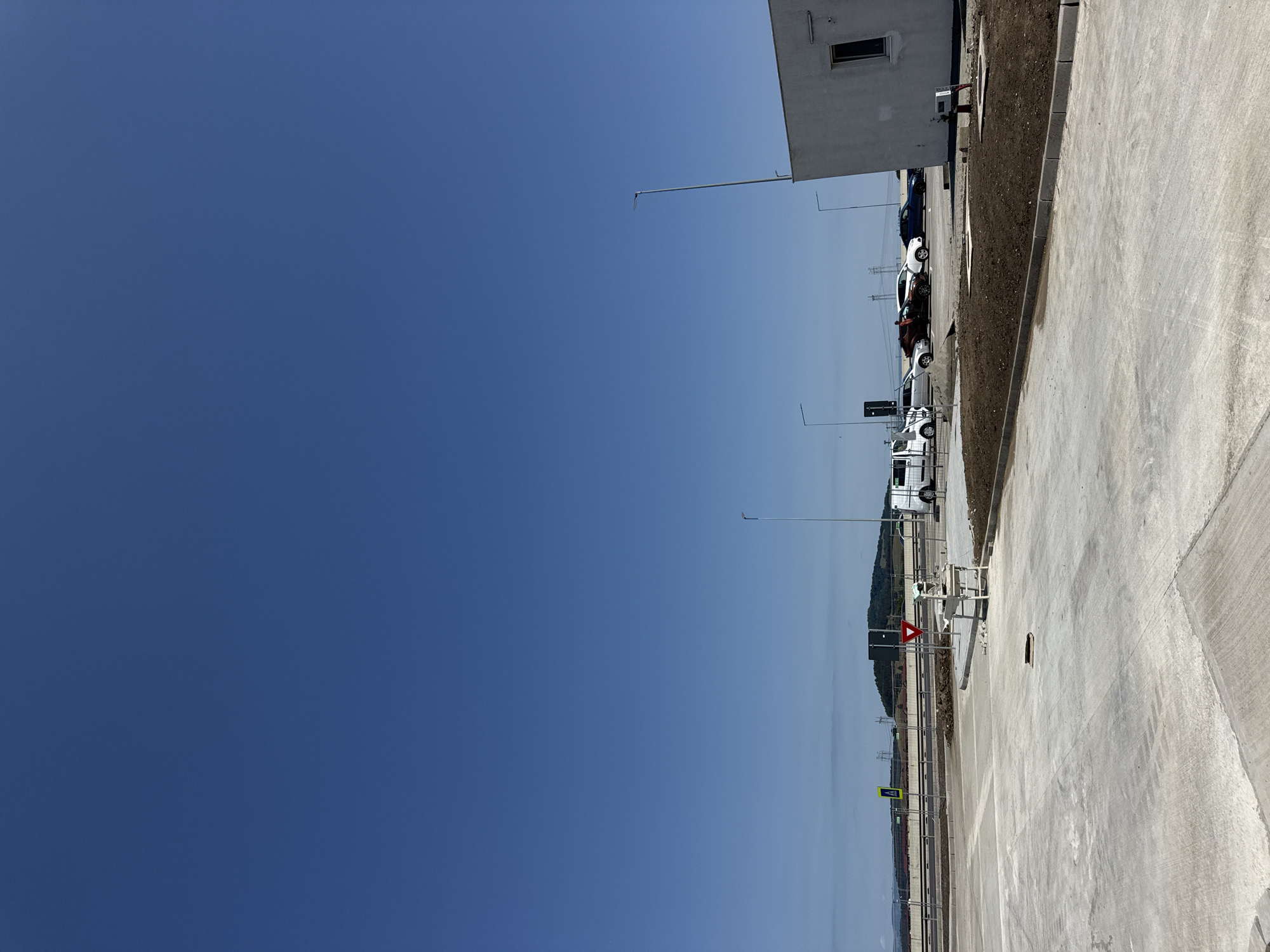 The image captures an outdoor scene in what appears to be a developing area in Sanpetru, Romania, on a clear day with a bright blue sky. In the foreground, a wide expanse of concrete paving dominates the view, suggesting a parking lot or construction site. To the right, a section of a white building with a single window is visible, with a small patch of dirt and some visible infrastructure at its base.
Further back, the scene transitions to a roadway or highway, bordered by a chain-link fence. Several vehicles are parked in a lot adjacent to this roadway. These include at least one white van, a dark-colored sedan, a white sedan, and a dark blue sedan. A person is seen interacting with the dark-colored sedan. A few streetlights are positioned throughout the scene. In the distance, a gently sloping hill or small mountain is visible under a hazy sky, with power lines and pylons dotting the landscape. Two traffic signs are also present: a red inverted triangle and a yellow diamond with a black pedestrian symbol. The ground is a mix of concrete, dirt, and some sparse vegetation.