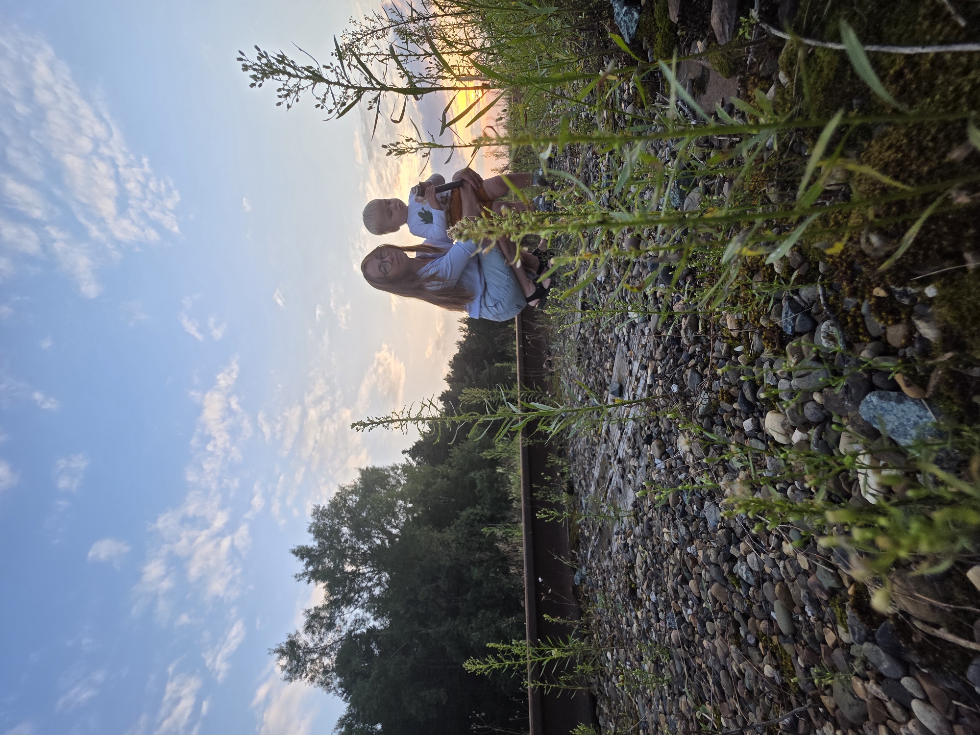 A woman kneels on a rocky surface, holding a young child. The woman has long, dark hair and wears glasses and a light blue shirt. The child, with blonde hair, is held in her arms and appears to be wearing a white shirt with a leaf emblem. They are positioned in the foreground, with lush green vegetation growing around them. Behind them, a forest of dark green trees stretches across the landscape. The sky above is a pale blue, with scattered white clouds, and the setting sun casts a warm glow, hinting at a late afternoon or early evening. The ground is covered in a mix of small stones and moss. A rusted metal rail runs horizontally through the scene, suggesting a location near a railway track. The overall mood is peaceful and intimate.