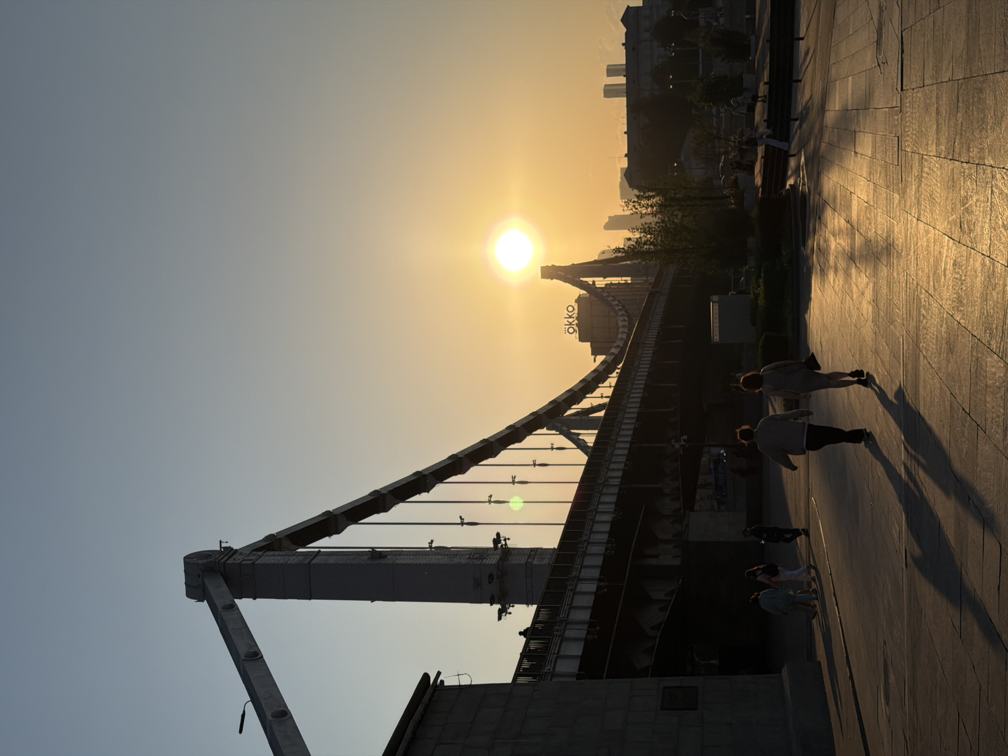 The image depicts a sunset view of a bridge, possibly a pedestrian bridge, in a city. The sky transitions from a light blue to a golden yellow where the sun is setting. The bridge is a dark silhouette against the bright sky. People are walking along the bridge and the adjacent walkway. The walkway is made of stone or concrete pavers, and the setting sun reflects off of its surface. In the background are city buildings. There is also a sign that says "okko".