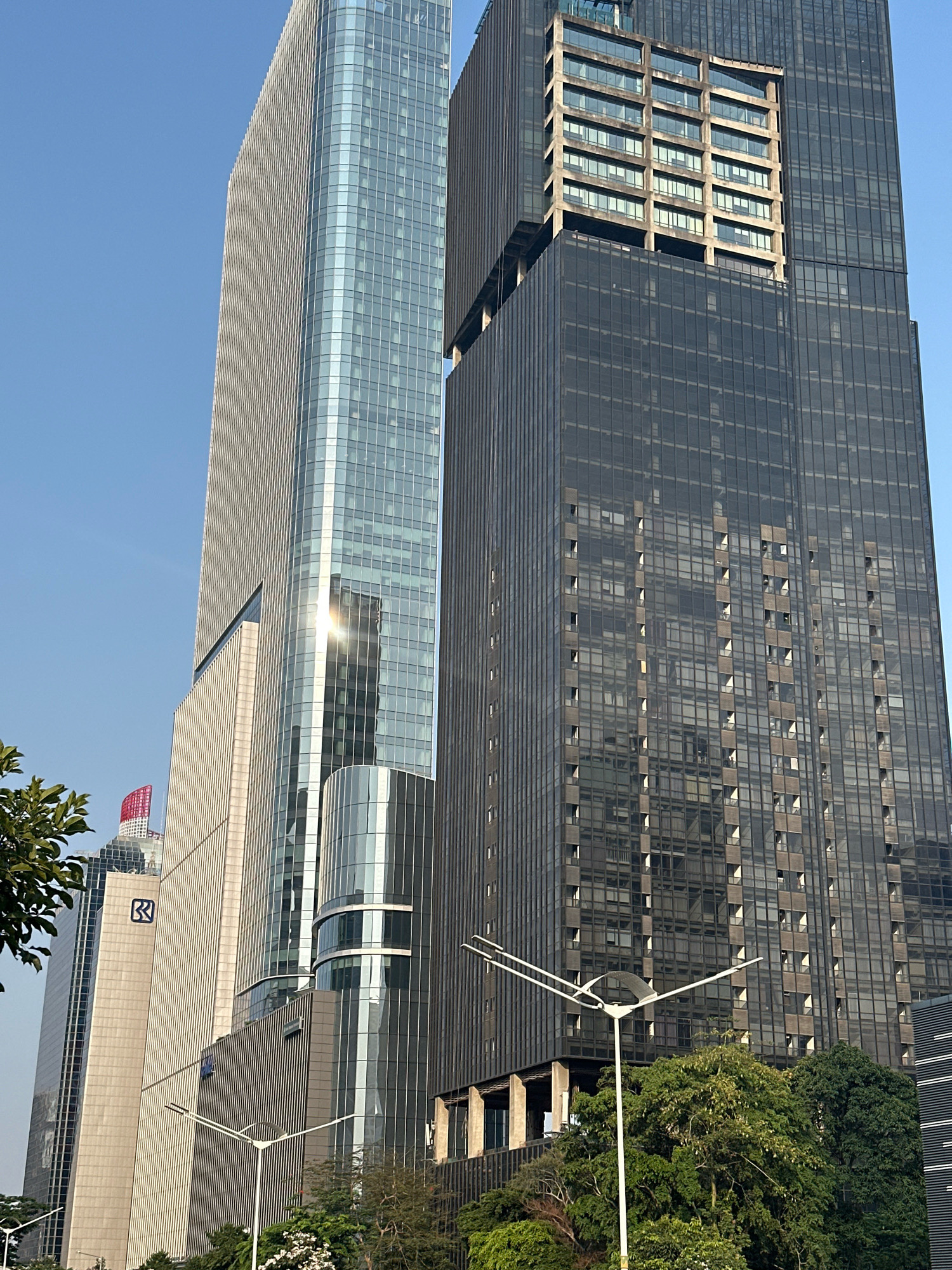 This is a low-angle, full-shot photograph of several modern skyscrapers in a city. The skyscrapers are primarily made of glass and steel, with some featuring concrete elements. The building on the left is tall and slender with a light-colored facade and reflective glass windows. The building in the center-right is dark and appears taller, with a facade of dark-tinted glass reflecting the sky and surrounding buildings. A portion of this building has a more industrial, concrete structure with many windows. To the far right, another dark glass skyscraper is partially visible. In the foreground, lush green trees and foliage obscure the lower portions of the buildings. A white streetlamp with a distinctive curved design stands prominently in the center foreground. The sky is clear and a bright blue. The scene appears to be in an urban business district, possibly in South Jakarta, Indonesia, given the architectural style and the presence of foliage common in tropical cities. No people are visible in the image.
