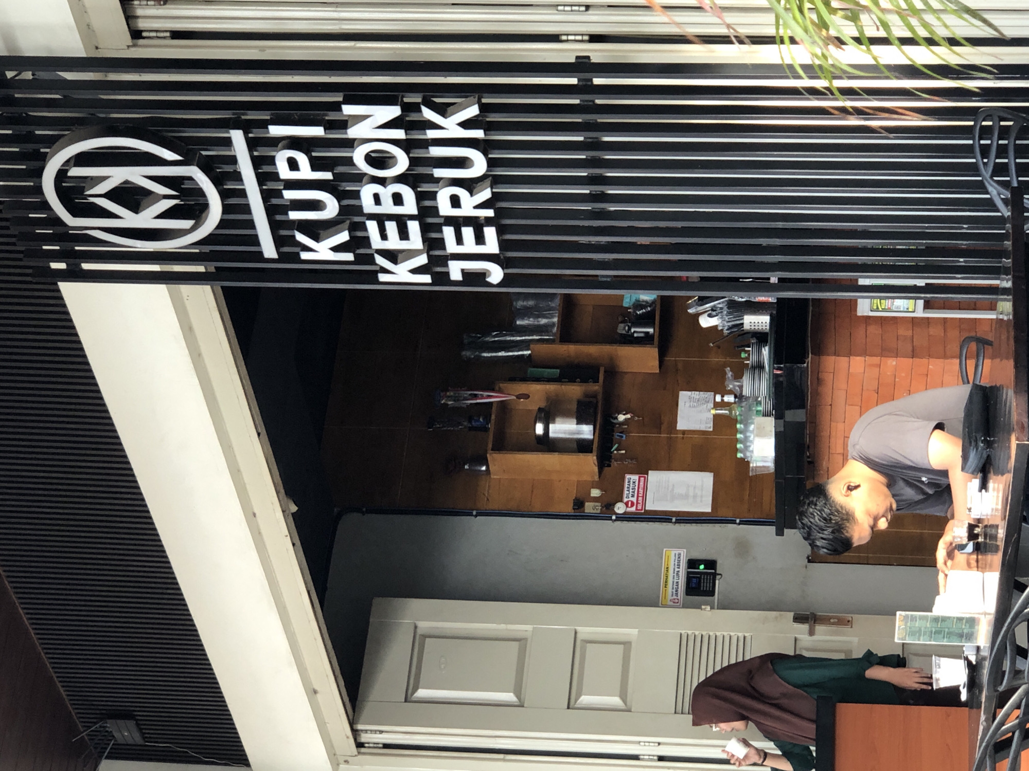 This media file depicts the exterior of a cafe named "KOPI KEBON JERUK". The sign, in white lettering against a black metal slatted background, is prominently displayed. The scene is set in what appears to be a commercial area, possibly during daylight hours, though the exact time is not discernible.
In the foreground, a man wearing a grey t-shirt is engaged in an activity at a counter, likely serving customers or preparing items. He is looking down, seemingly focused on his task. Behind him, a woman wearing a green outfit and a brown hijab is also present, holding a white object.
Various objects are visible on shelves and countertops within the cafe, including bottles, containers, and what appear to be service equipment. A white door is visible in the background, as well as brickwork on the wall. A plant with green leaves is hanging from above, adding a touch of natural element to the scene.
There are several signs with text visible. One sign, in red and white, reads "DILARANG MASUK! DALAM KARYAWAN" which translates to "NO ENTRY! EMPLOYEES ONLY". Another sign, in yellow and black, states "PERHATIAN! JANGAN LUPA ABSENSI" which translates to "ATTENTION! DON'T FORGET TO CLOCK IN". A digital display near this sign shows "17:32".
The overall impression is that of a busy cafe environment.