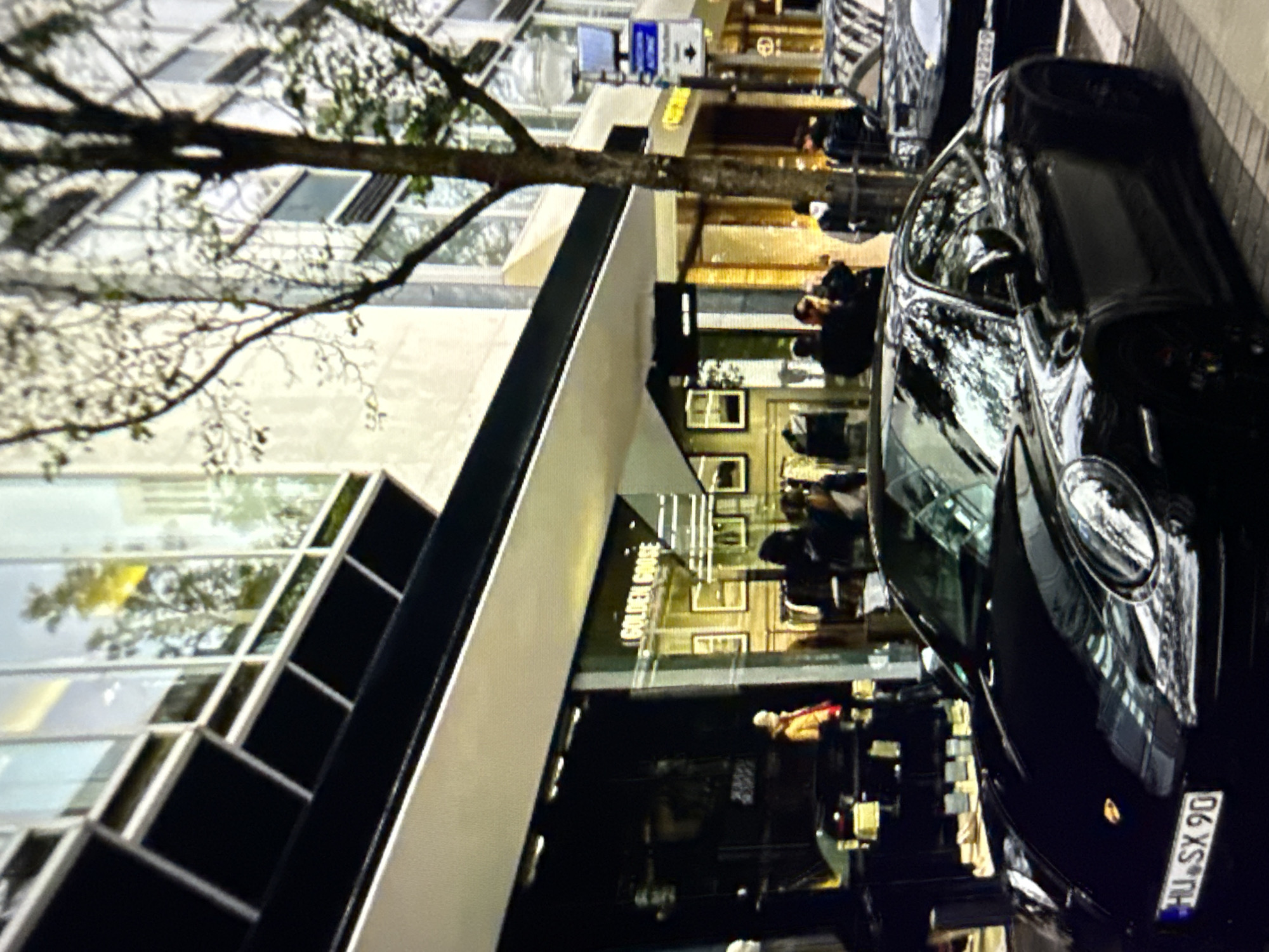 A street-level urban scene is depicted under daylight conditions, showcasing a prominent black Porsche 911 parked at the curb in the foreground. The Porsche displays a German license plate reading "HU SX 90". Another dark-colored luxury vehicle, possibly a Porsche, is parked directly behind it, with a partially visible license plate showing "U 2019", consistent with a German format. The vehicles are positioned on a cobblestone-patterned sidewalk next to a road.
On the left, behind the first Porsche, is the storefront of a "GOLDEN GOOSE" store, identified by its illuminated sign. Reflections of interior lighting and items are visible in its large glass windows. A person wearing a bright yellow/orange jacket is visible inside the store. Further inside, framed pictures adorn a wall.
The buildings on both sides of the street are multi-story structures with numerous large windows. Reflections of trees and the sky are visible in the windows of the upper floors and on the surfaces of the parked cars. A bare-branched tree stands on the right side of the image, partially obscuring the background building and a street pole with various signs. Several indistinct figures of people are visible walking on the sidewalks or reflected in shop windows, suggesting pedestrian activity. The overall environment is an upscale commercial street.