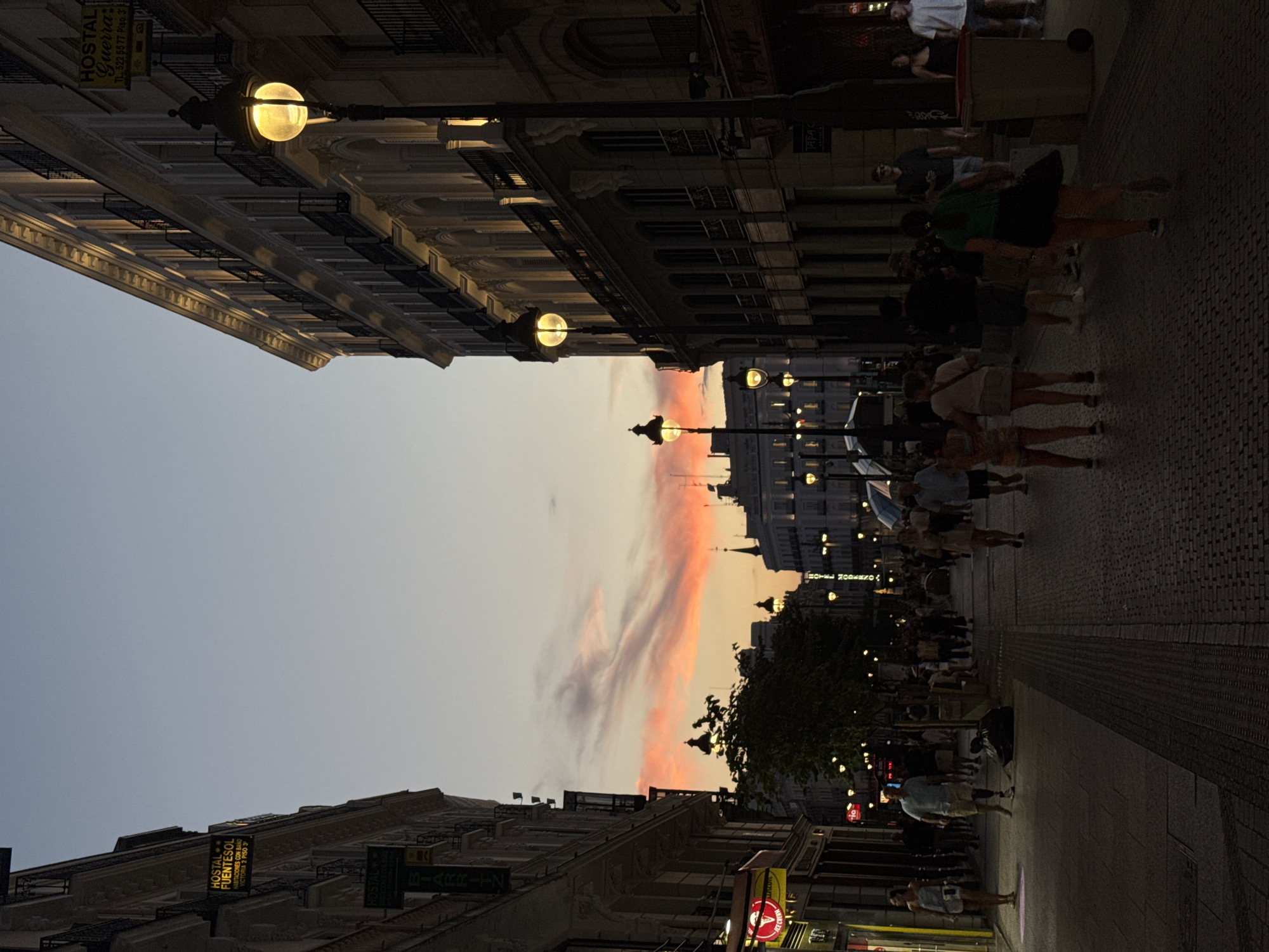 This image captures a street scene in Madrid, Spain, during twilight. The sky displays a gradient of blues and pinks, with streaks of orange from the setting sun, indicating it is either sunrise or sunset. The street is paved with cobblestones and lined with buildings on both sides. The buildings are multi-story, with ornate architecture, balconies, and numerous windows. Streetlights, some illuminated with a warm, yellow glow, are visible along the sidewalk.
A diverse group of people are walking along the street, some in groups and others individually. They are dressed in casual summer attire. One person appears to be walking with a prosthetic leg. There are shops and signs visible on the buildings, including what appears to be a "Hostal Fuentesol" and another hostel sign. A trash bin is present on the right side of the street. Trees are also scattered along the pedestrian walkway. The overall atmosphere suggests a lively urban environment at the end of the day.
