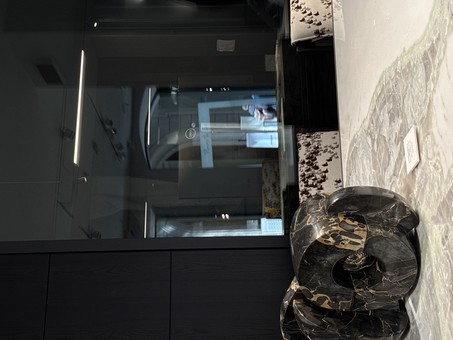 This image captures an interior scene, likely within a modern showroom, high-end office, or design studio in Milan, Italy. The photograph is presented on its side, with the primary elements arranged horizontally across the frame.
To the left, a large, dark, reflective glass panel or partition dominates, mirroring various elements within and beyond the space. Visible reflections include bright, vertical strip lights, and a digital display featuring the "DELL" logo along with partially legible text such as "Meeting Room." Blurred reflections of several individuals are also discernible, suggesting people are moving or standing nearby. Below this reflective surface, a dark-toned wooden or paneled wall adds to the sleek aesthetic.
To the right, a striking surface of light-colored marble or natural stone serves as a display area. This slab exhibits elegant grey and greenish veining patterns. Resting upon this marble is a substantial, abstract sculpture crafted from black marble, intricately veined with rich gold and tan patterns, forming smooth, complex curves.
Behind the sculpture and on the marble surface, several upholstered display blocks or small stools are visible. They are covered in a textured, light-colored fabric with a repetitive, dark speckled pattern. A small white card sits on the marble next to the sculpture, with visible Italian text: "MATERIALI E DESIGN DI..." followed by "Pietra Naturale," indicating a focus on natural stone materials and design.
The overall atmosphere is sophisticated and contemporary, with a strong emphasis on high-quality materials and clean lines. No specific activity beyond casual viewing or passing by is evident, and the lighting suggests an indoor setting with artificial illumination.