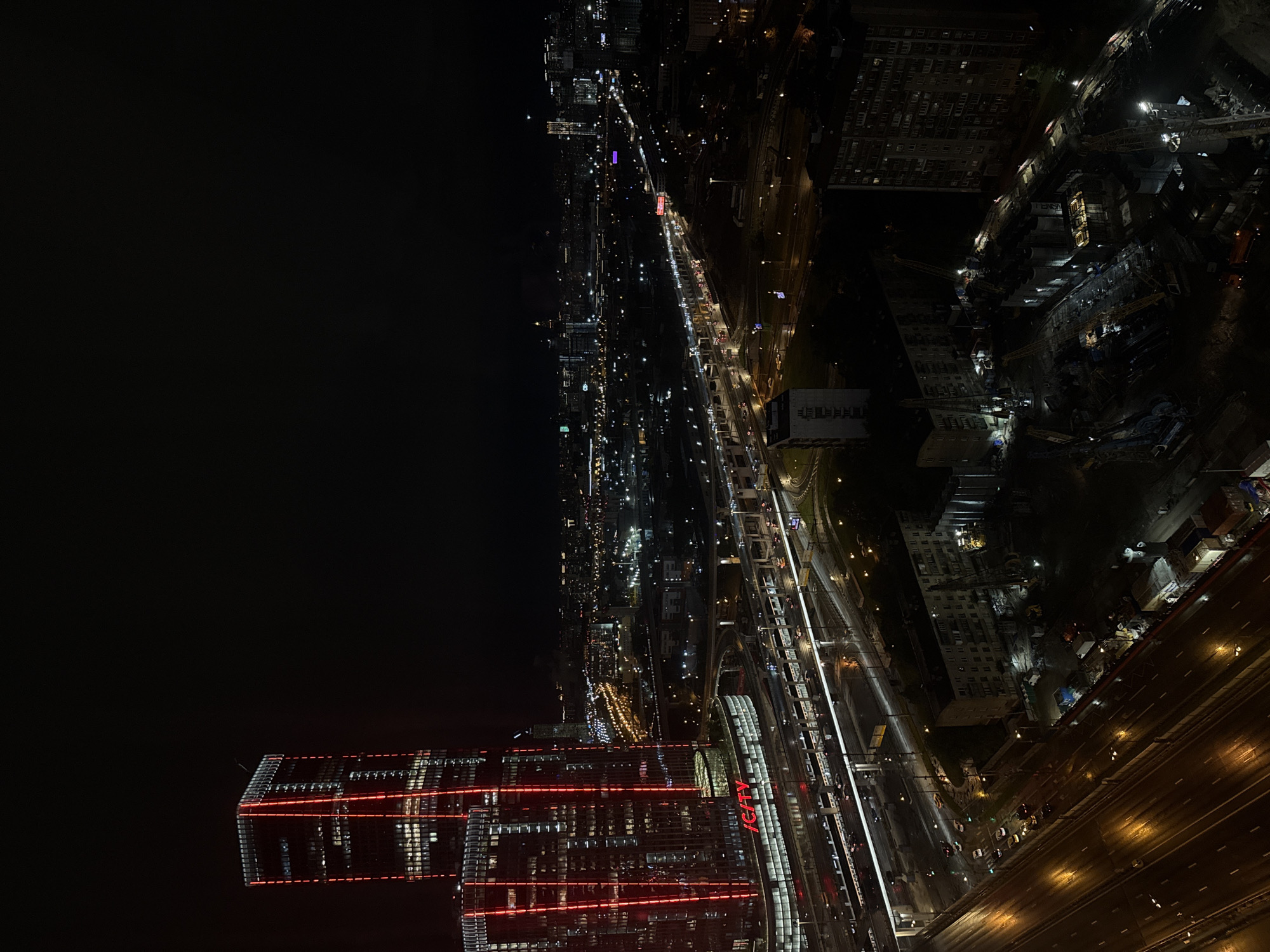 Here is a detailed description of the image:
The image shows a nighttime view of a city, possibly taken from a high vantage point like a skyscraper. The scene is dominated by city lights illuminating buildings, streets, and infrastructure.
In the foreground are illuminated skyscrapers with red horizontal lines. The streets below show light trails of moving vehicles. Alongside is a railroad track, brightly illuminated. The middle ground shows a grid of city streets and residential buildings. Construction sites are visible with construction cranes and active lighting. The city extends to the horizon, with diminishing light and detail.
The environment is clearly urban, with a focus on nighttime illumination and the structures of modern city life.