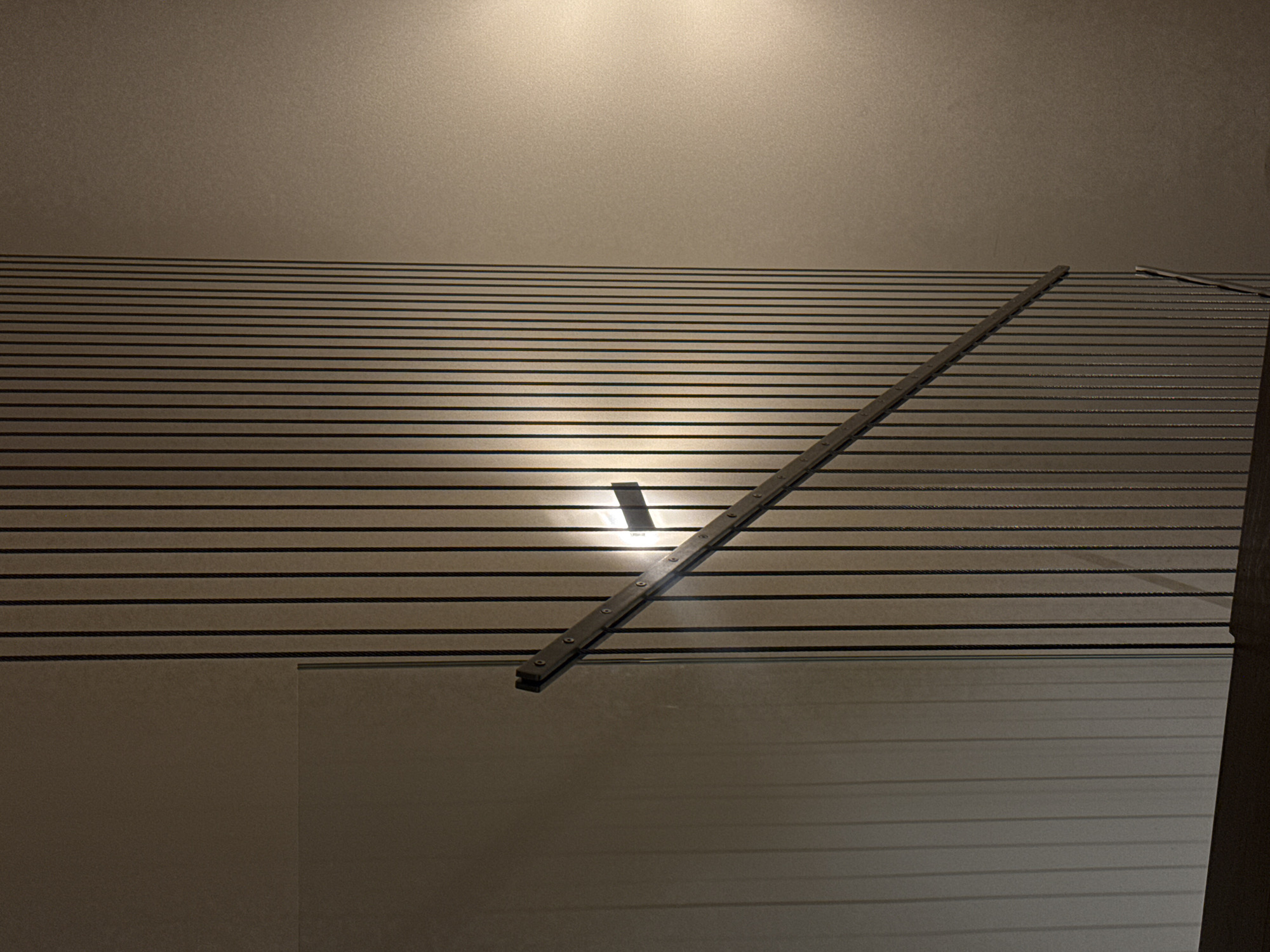 The image depicts a section of the ceiling and possibly a wall in an interior space. The ceiling is made of horizontal slats. A metal bar with equidistant holes runs diagonally across the ceiling. Below the ceiling, a reflective surface is visible, likely a wall. There is no indication of the image being in Tel-Aviv, Israel. There are no people present in the image.