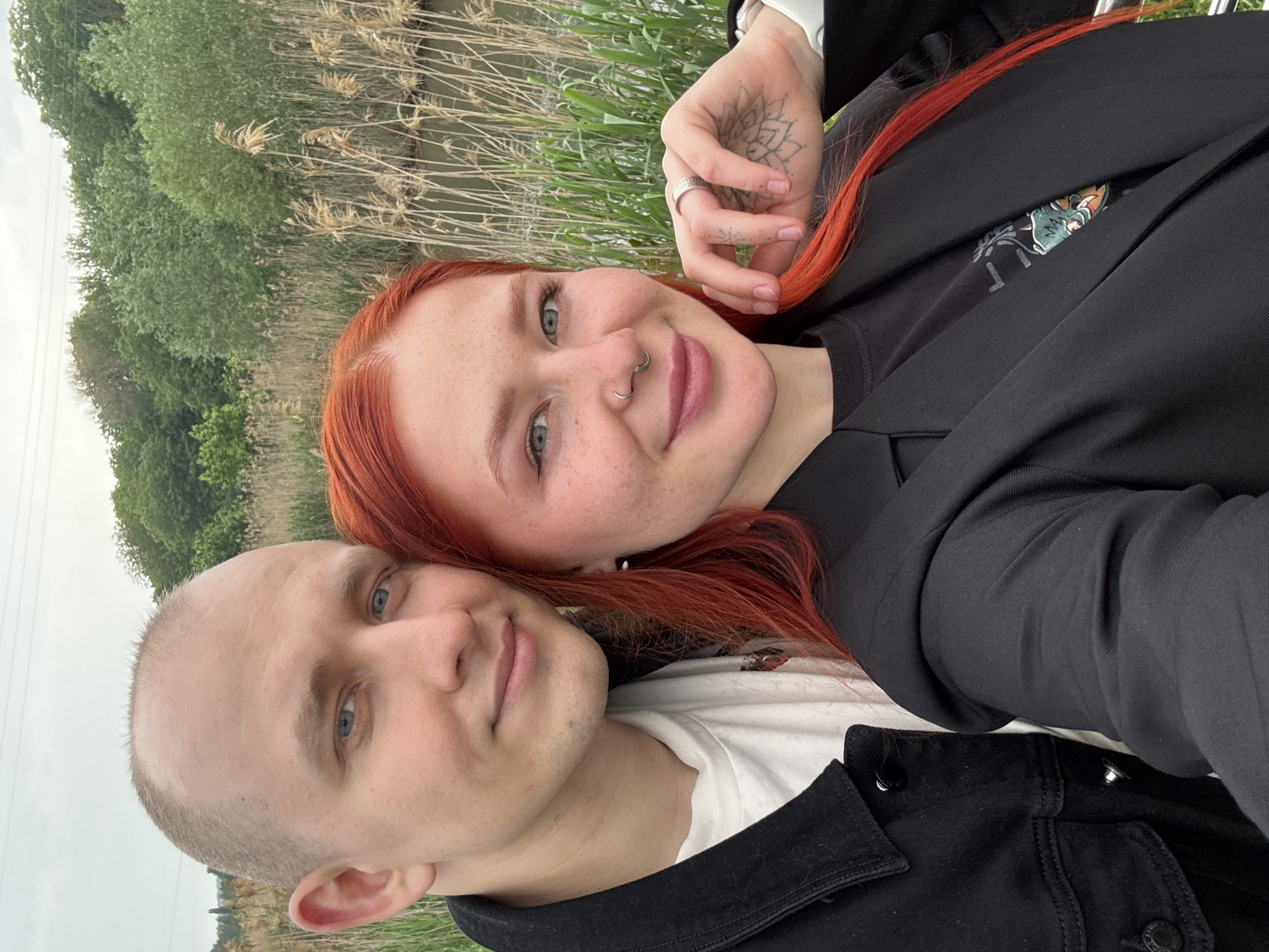 Two individuals, a man and a woman, are pictured in a close-up selfie outdoors. The woman has red hair and a nose piercing. She is wearing a black jacket over a black shirt. Her left hand is visible, adorned with a ring and a tattoo. The man is bald and is wearing a white shirt and a black jacket. Behind the individuals is a blurred background of greenery, appearing to be reeds along a body of water, potentially a river or lake. No definitive landmarks are visible to confirm the location as Rostov-on-Don, Russia.