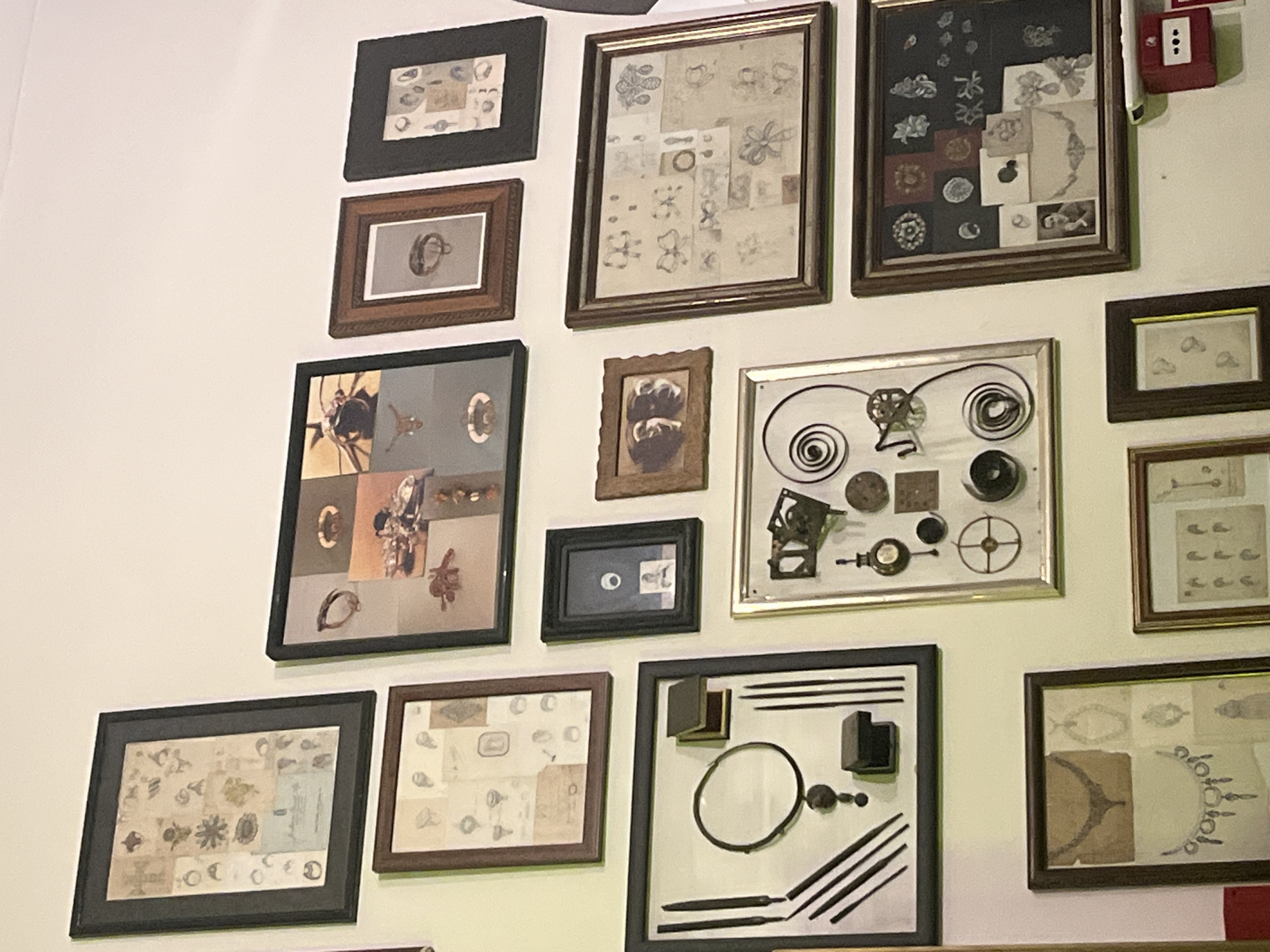 The image shows a wall covered in framed art pieces. The frames are various sizes and colors (black, brown, silver). The content of the frames seems to be sketches, photographs, and arrangements of small mechanical parts. To the upper right, there is a red fire alarm pull station on the wall.
The location is Lisbon, Portugal.