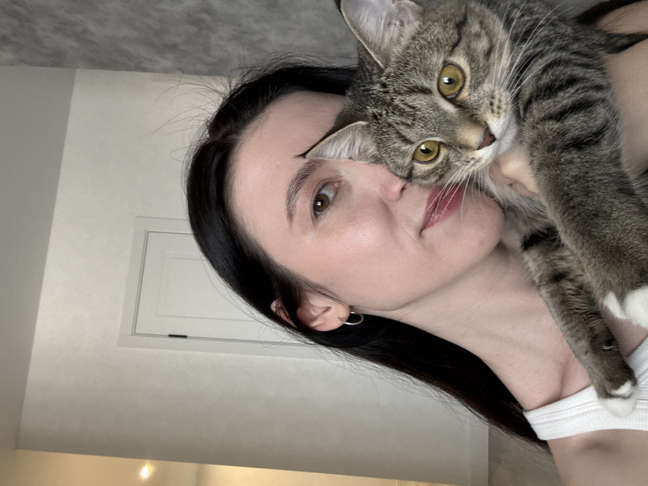 A female individual holds a tabby cat in an interior residential setting in Kazan, Russia. The woman, with long dark hair and fair skin, wears a white ribbed tank top and a silver hoop earring in her left ear. She looks towards the camera with a subtle smile, her right side partially obscured by the cat. The cat, a grey-brown tabby with distinct stripes, white paws, and amber eyes, is held close to the woman's chest and also gazes in the direction of the camera. The background features light-colored, possibly textured, walls, including a white door with a dark vertical handle on the left, and a section of grey textured wall on the right. A light source is visible in the upper left background.