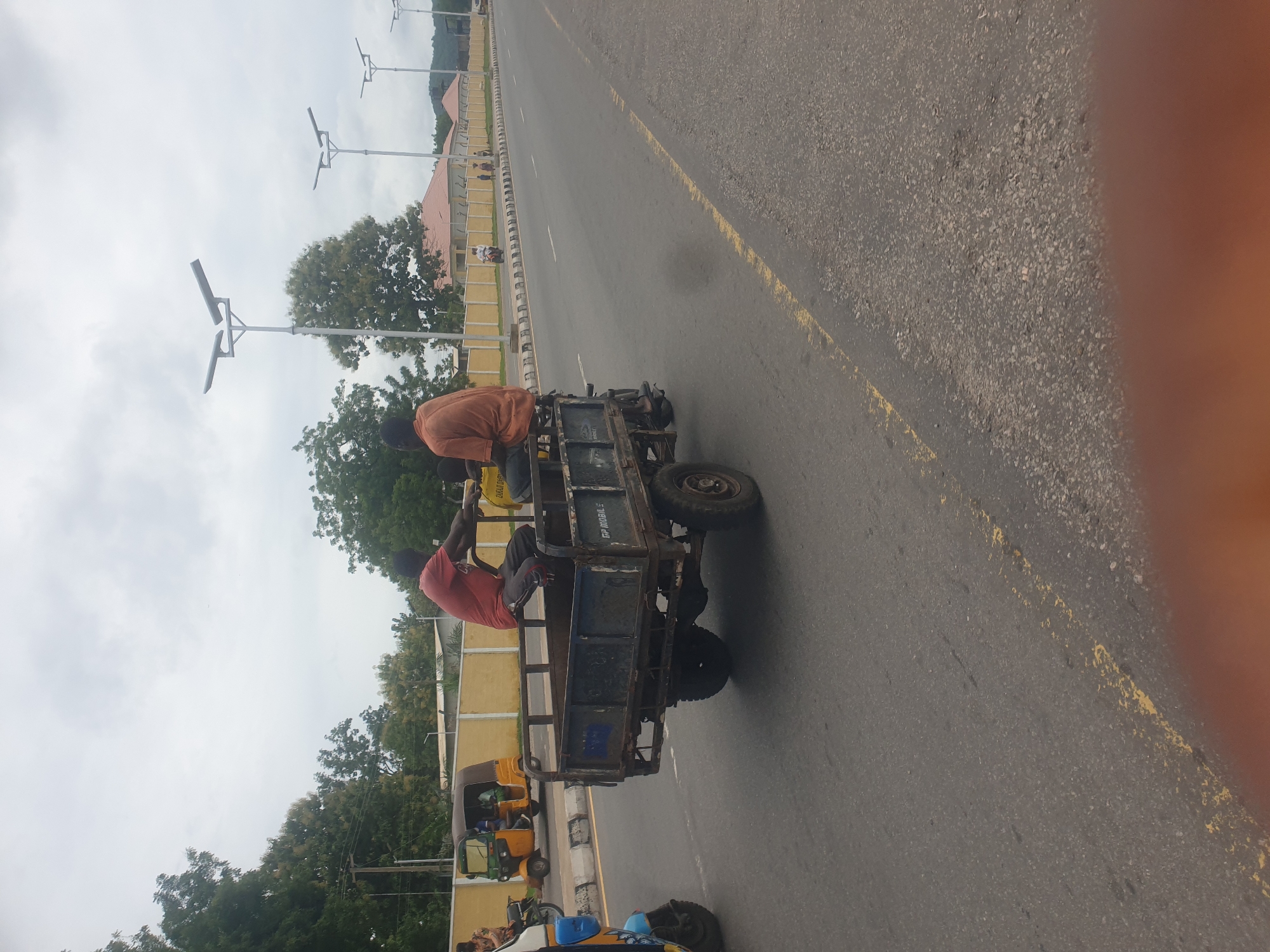 On a gray asphalt road with yellow lines, two men are riding in the back of a blue, three-wheeled motor vehicle with a cargo bed. The man in the front wears an orange shirt, while the man in the back is in red. The vehicle is traveling on a road alongside a sidewalk and a low yellow and white wall. Beyond the wall are green trees and street lights. In the distance is a building with a red roof. Other three-wheeled vehicles can be seen parked along the sidewalk. An orange object is in the lower right corner. The location is Unknown, Unknown.